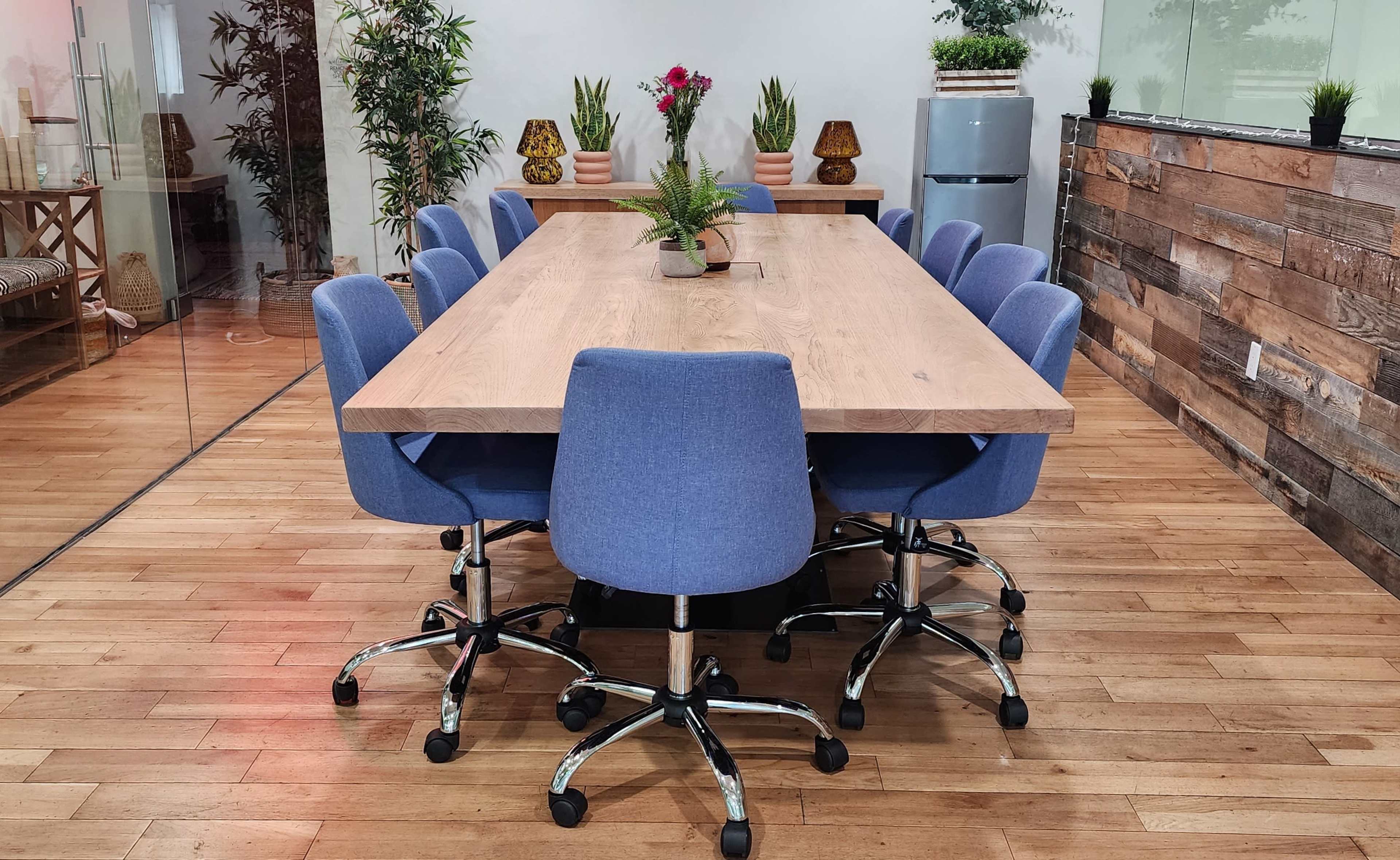 A long wooden conference table surrounded by eight blue upholstered chairs is set in a well-decorated meeting room with potted plants and a wooden accent wall.