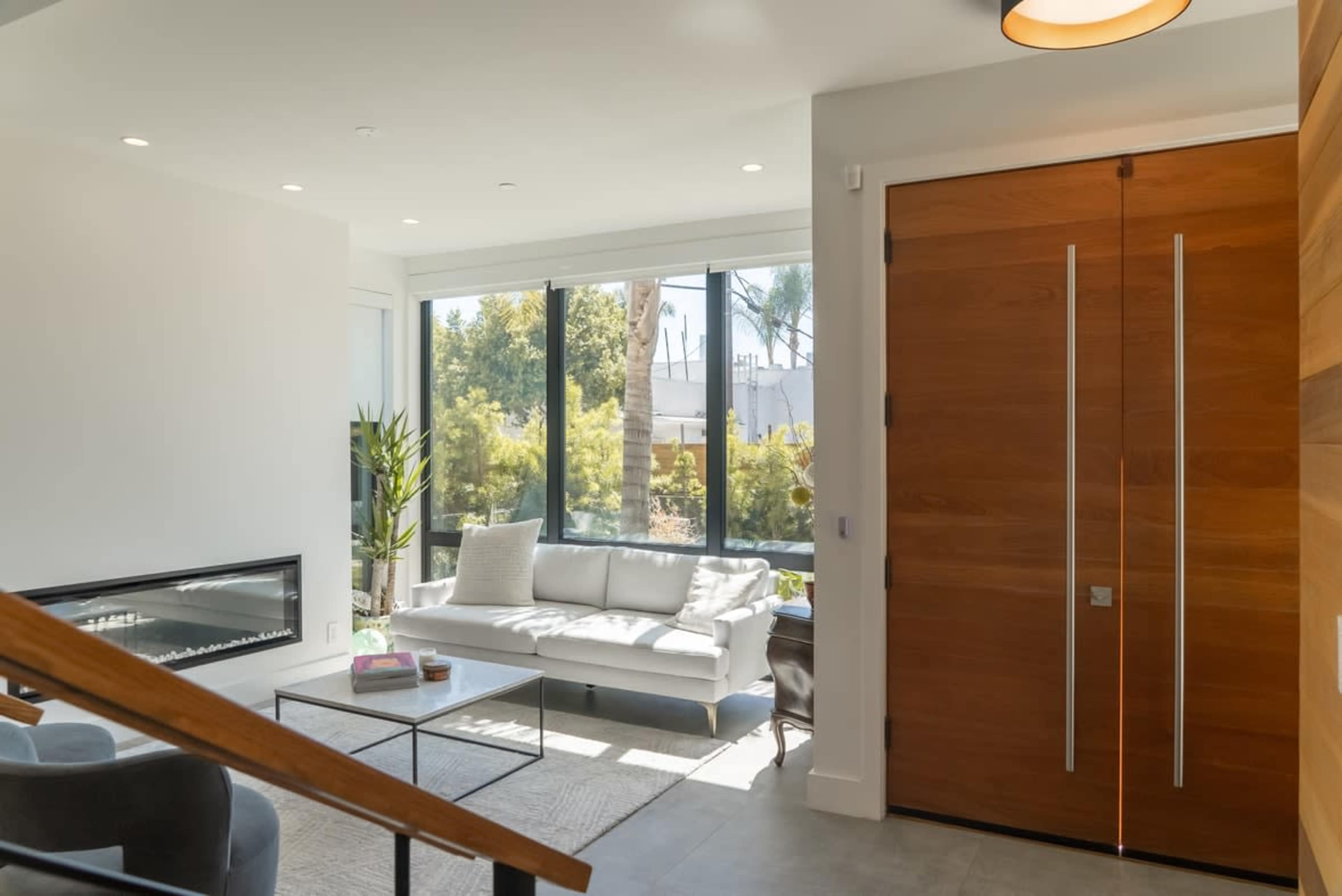 A modern entryway features a large wooden door, a white sofa, and a view of a garden through a large window.