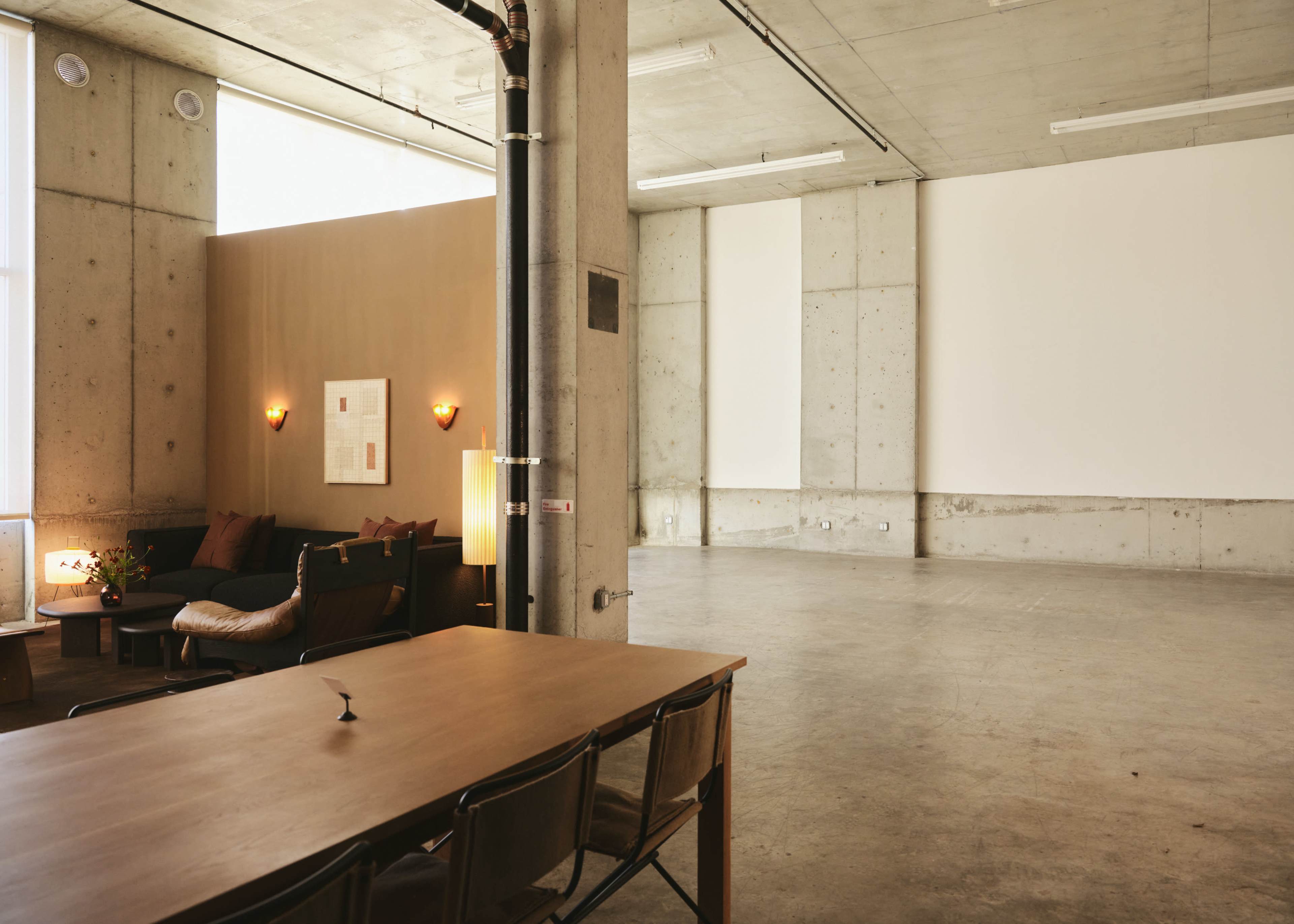 The image shows a spacious, industrial-style room with concrete walls, a seating area on one side, and an empty section suitable for events or presentations.
