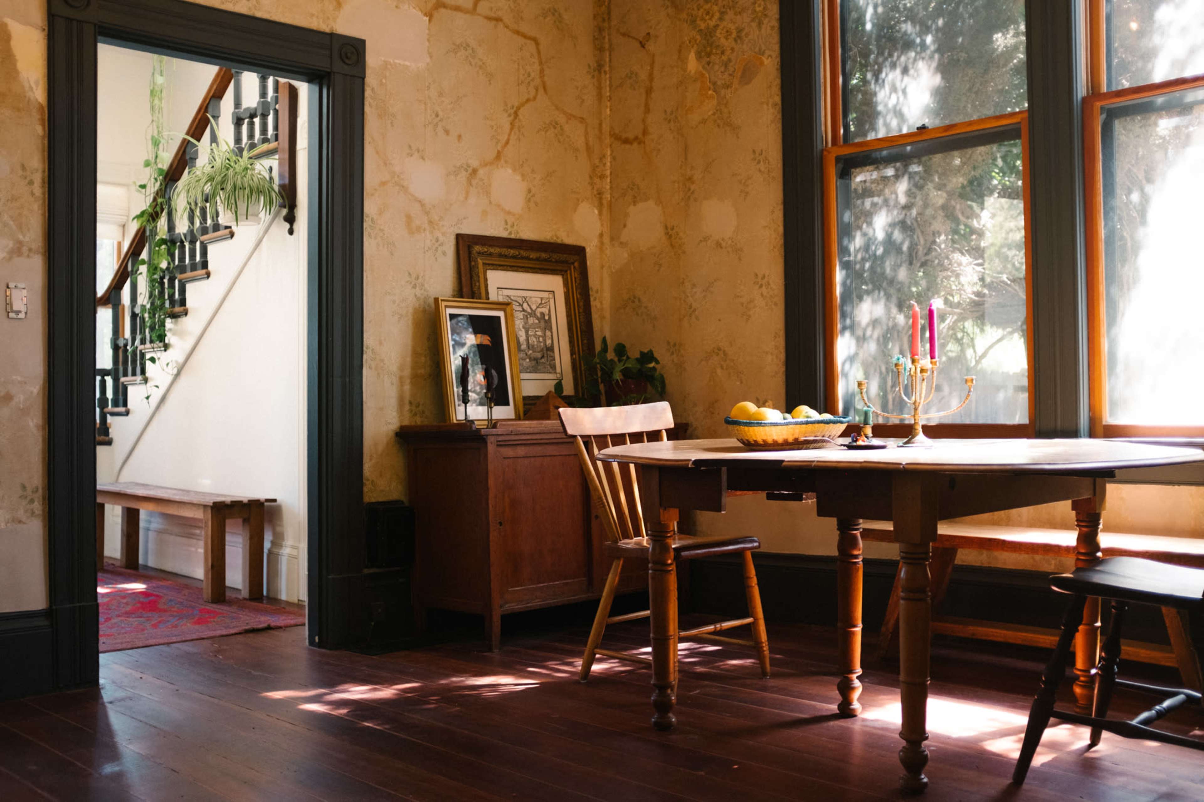 A wooden dining table with candles and fruit sits in a room featuring patterned wallpaper and a view of a staircase.