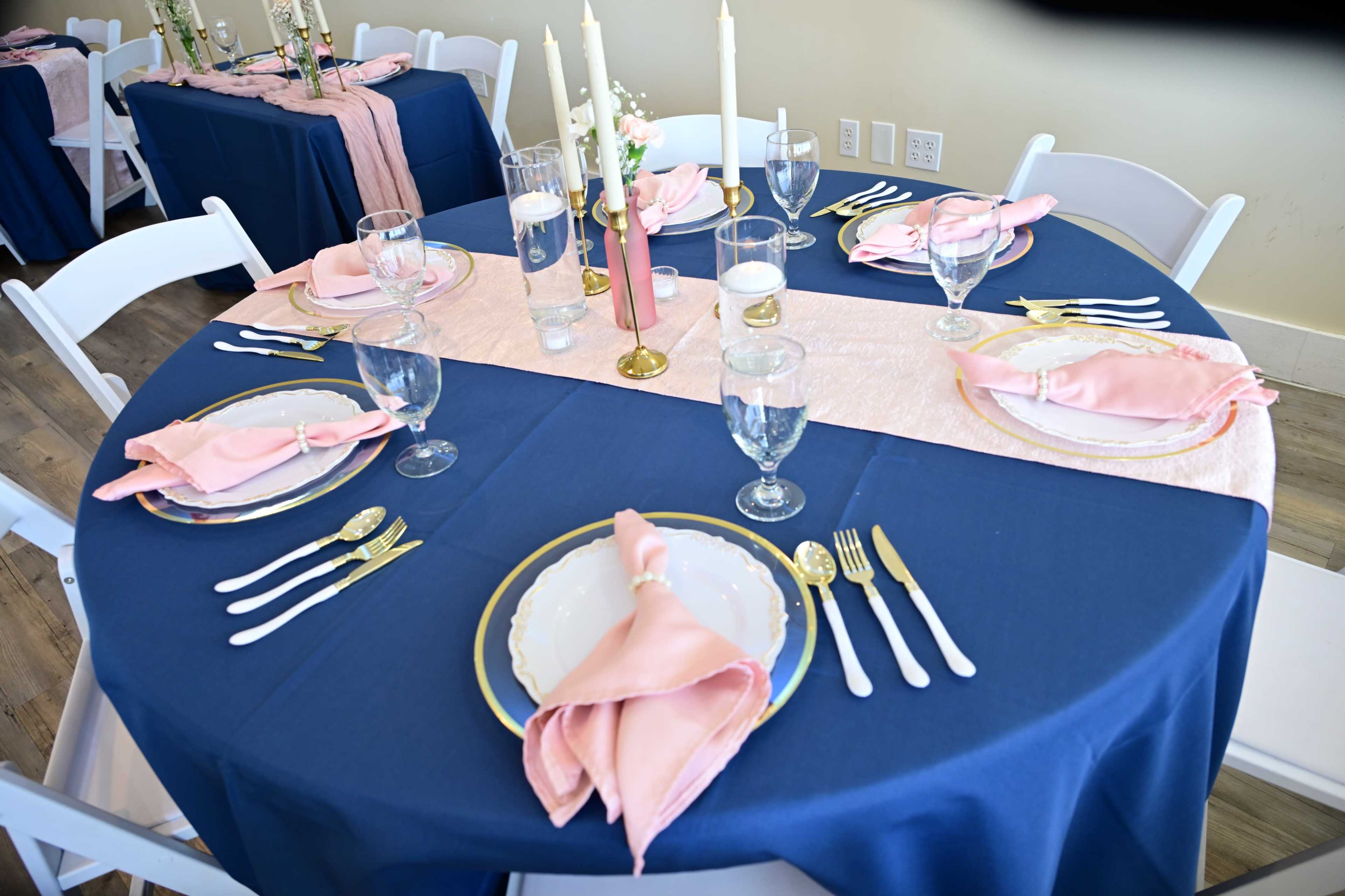 The image shows a beautifully set dining table with navy blue tablecloths, pink napkins, glassware, and gold-accented plates, arranged for an event.