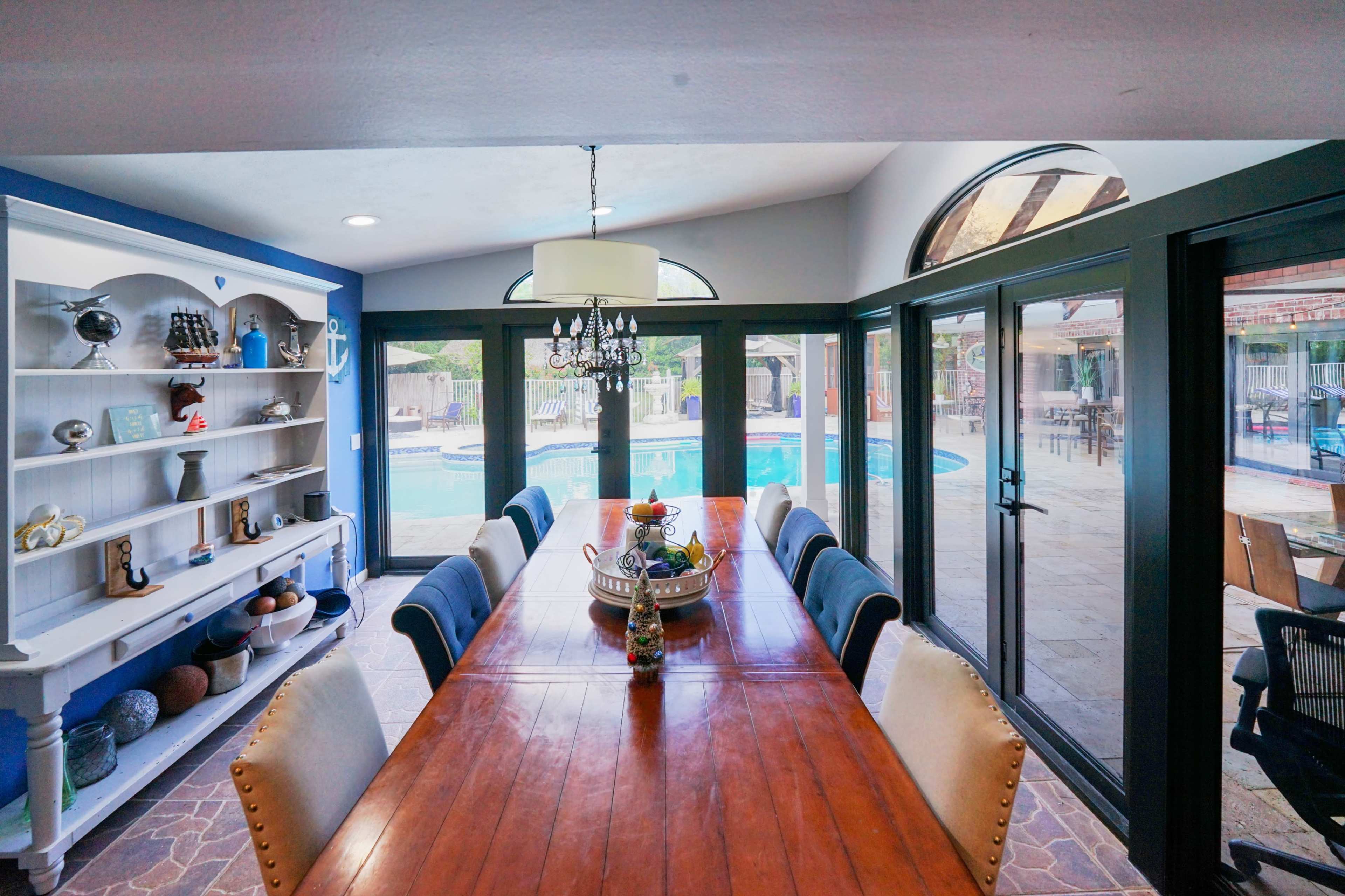 A long wooden dining table is set in a bright room with large windows overlooking a swimming pool area.