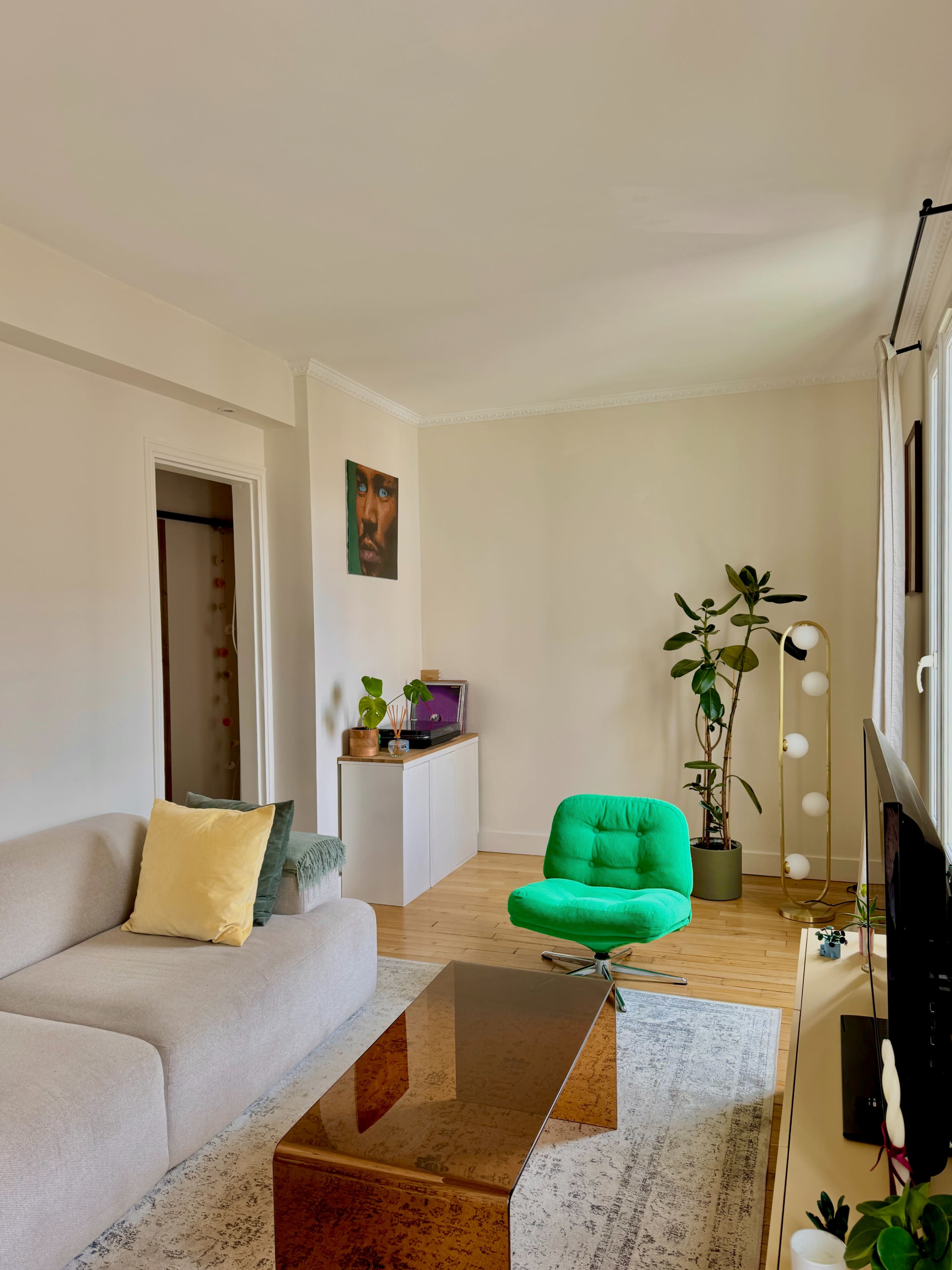 The living room features a light-colored sofa, a green lounge chair, a glass coffee table, and a potted plant, with a painting on the wall and natural light coming in through the windows.