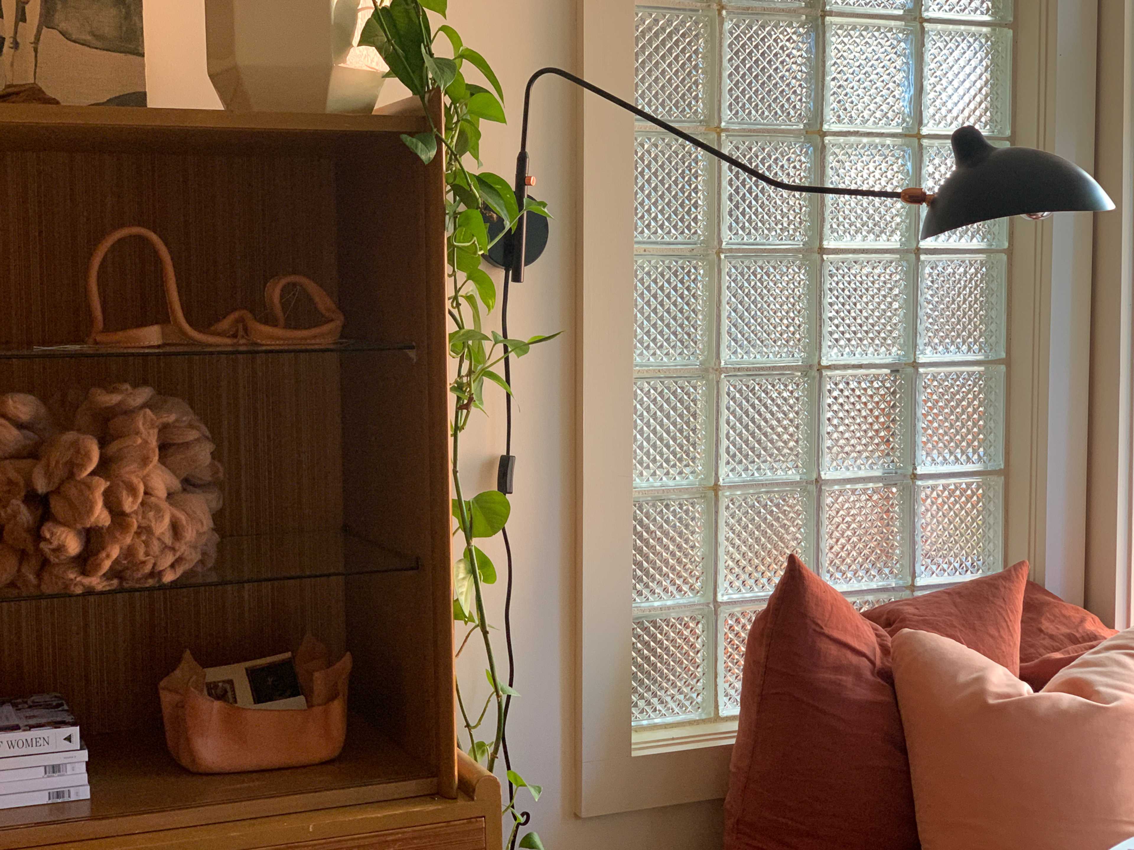 A mid-century modern bookshelf displays decorative items beside a glass-block window, with a floor lamp and soft cushions in the foreground.