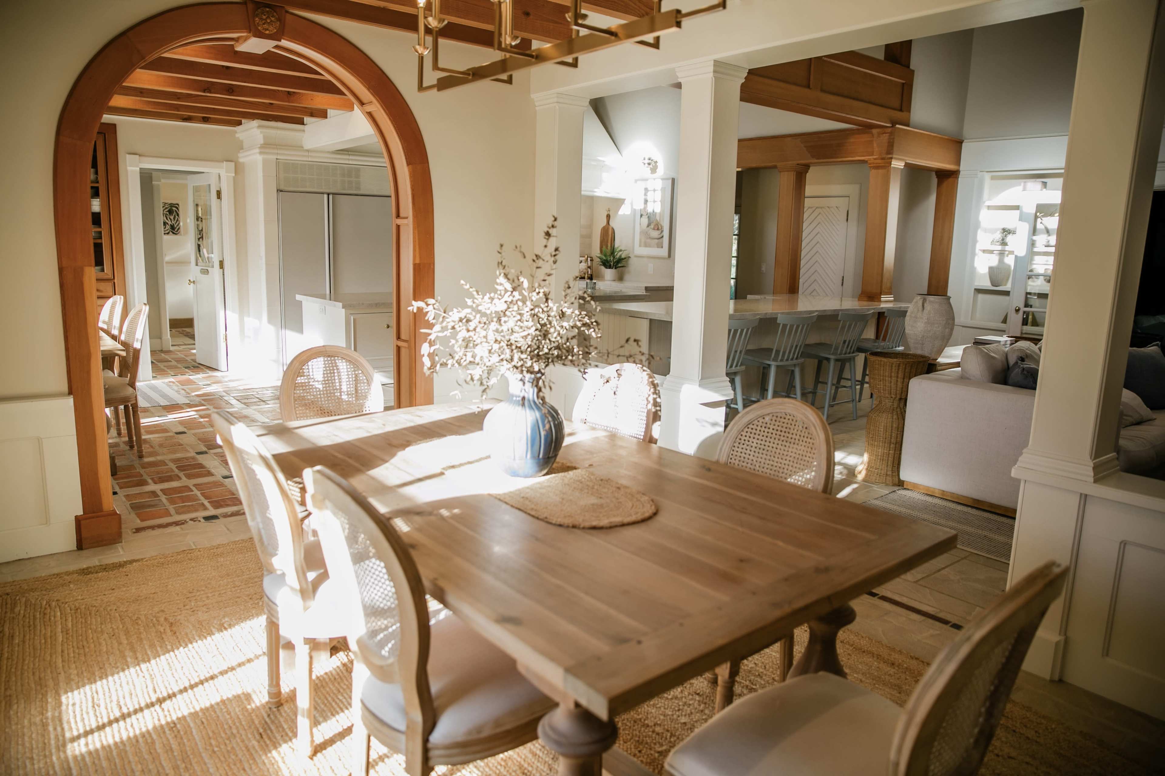 A dining area features a wooden table surrounded by light-colored chairs, adorned with a vase of flowers, with an open view into a bright kitchen and living space.