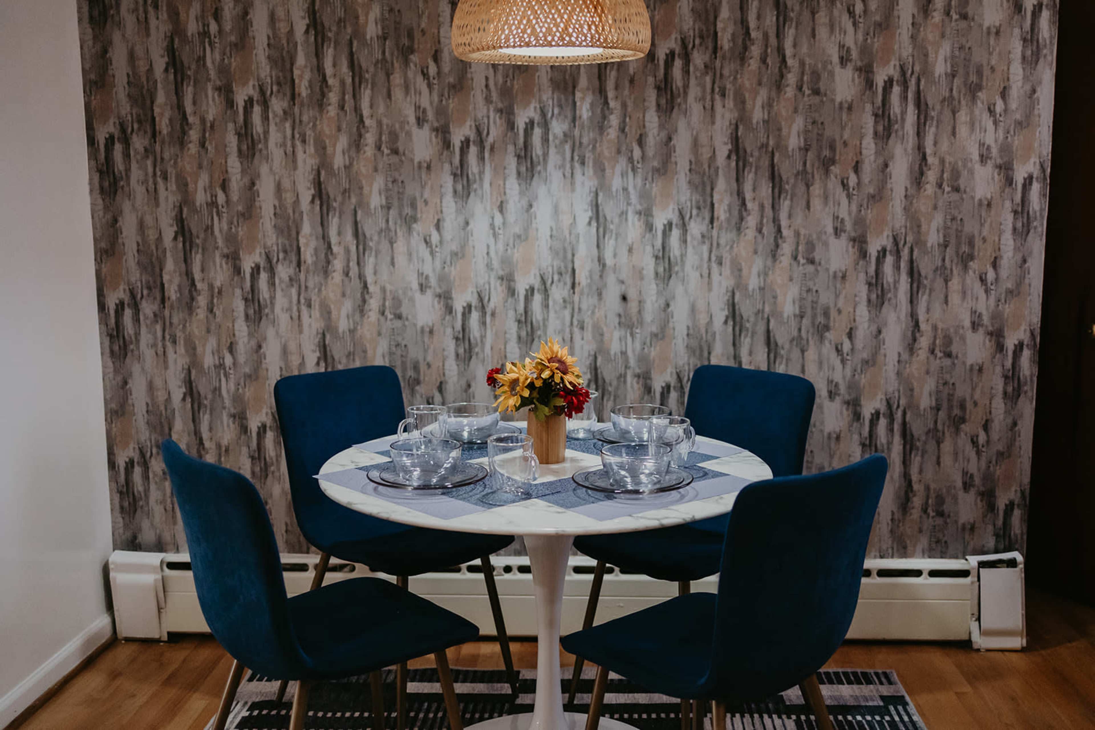 A round dining table with four blue chairs is set with clear glassware and a small vase of flowers, located against a textured wallpaper background.