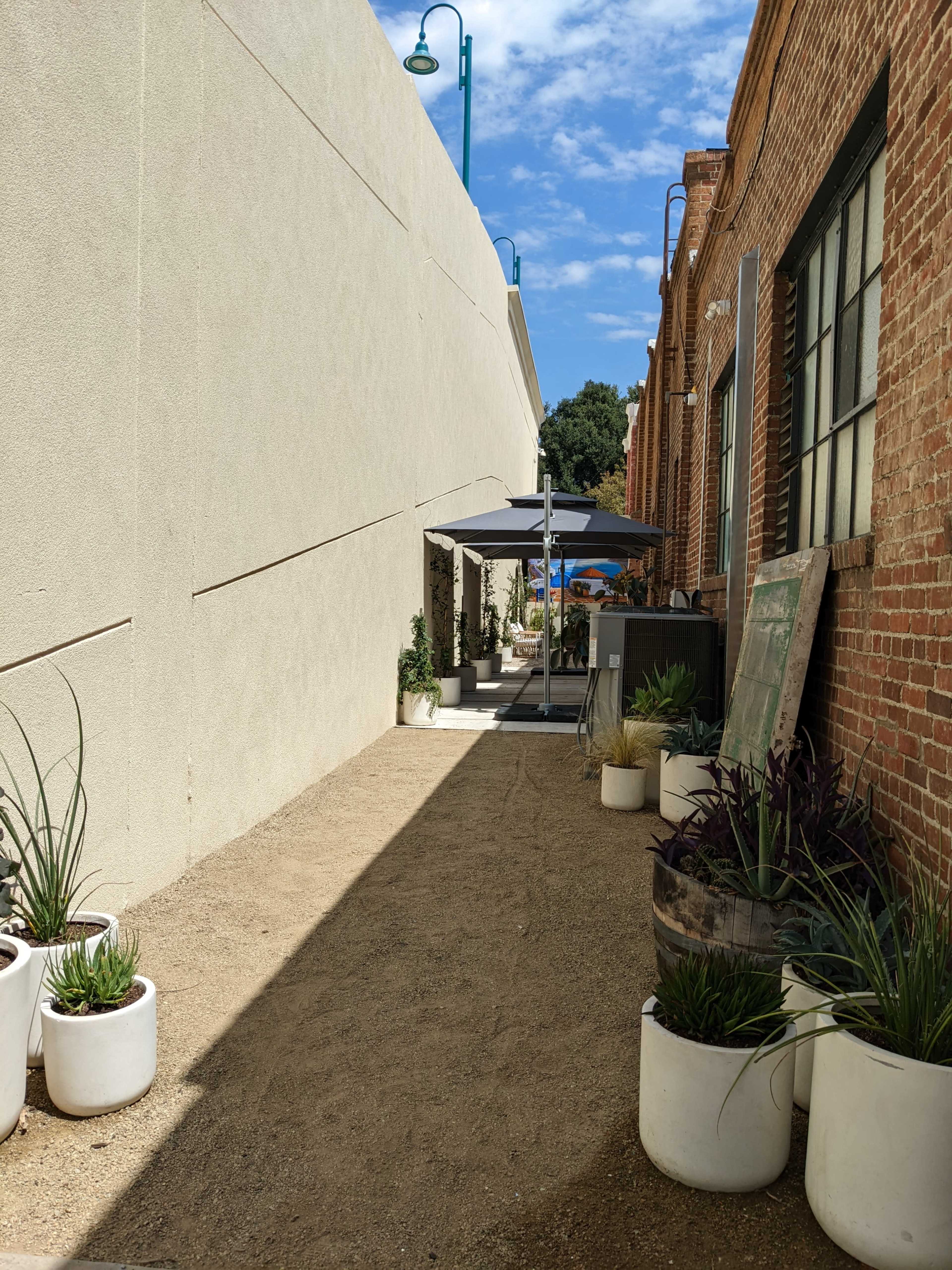 The image shows a narrow alleyway with potted plants on both sides, leading to a shaded area with an umbrella.