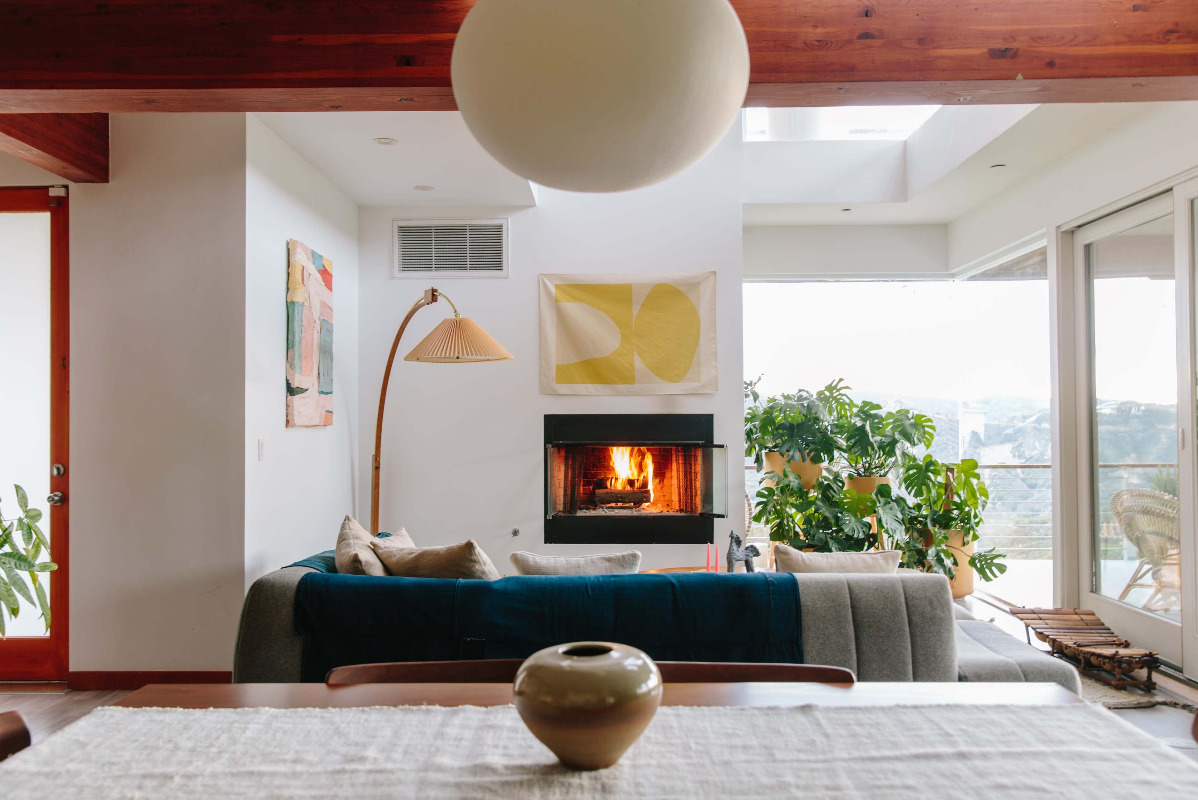 A spacious living room features a gray sofa, a floor lamp, a fireplace with a lit fire, and large windows with a view of greenery.