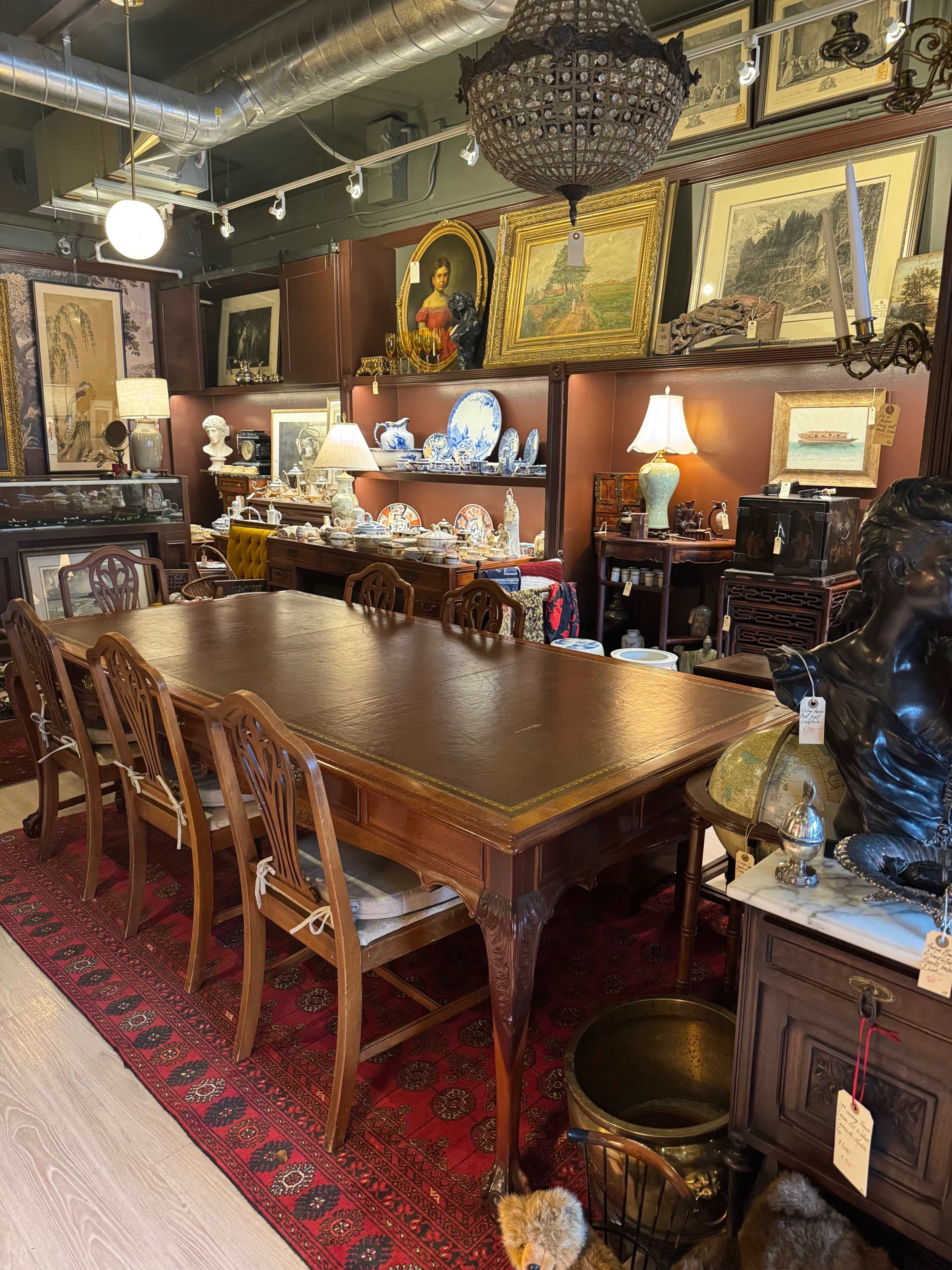 The image shows a vintage furniture store featuring a large wooden dining table surrounded by chairs, with various antiques and decorative items displayed on shelves and walls.