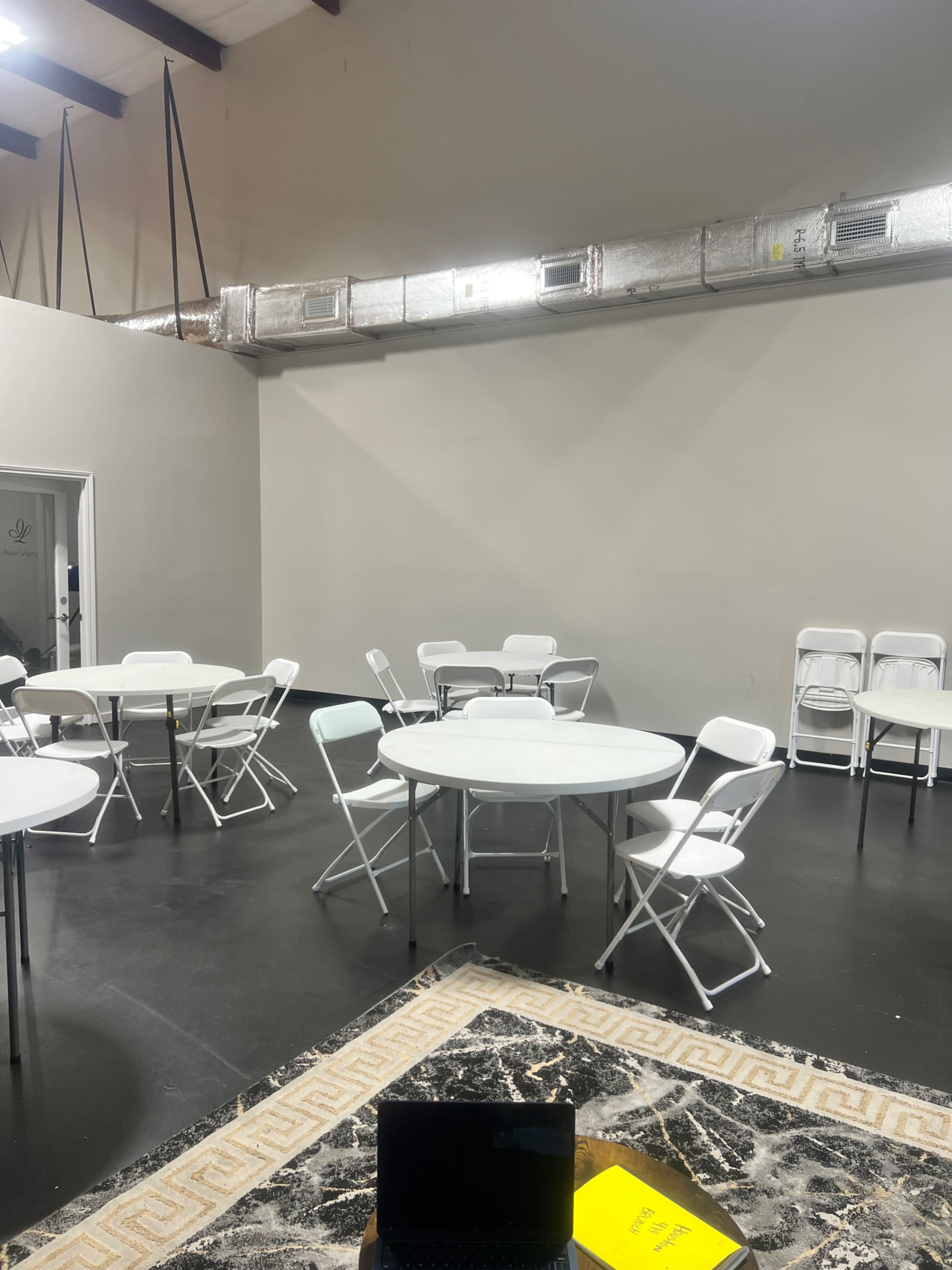 The image shows a spacious indoor area with white tables and chairs arranged around a central rug, while ductwork runs along the ceiling.