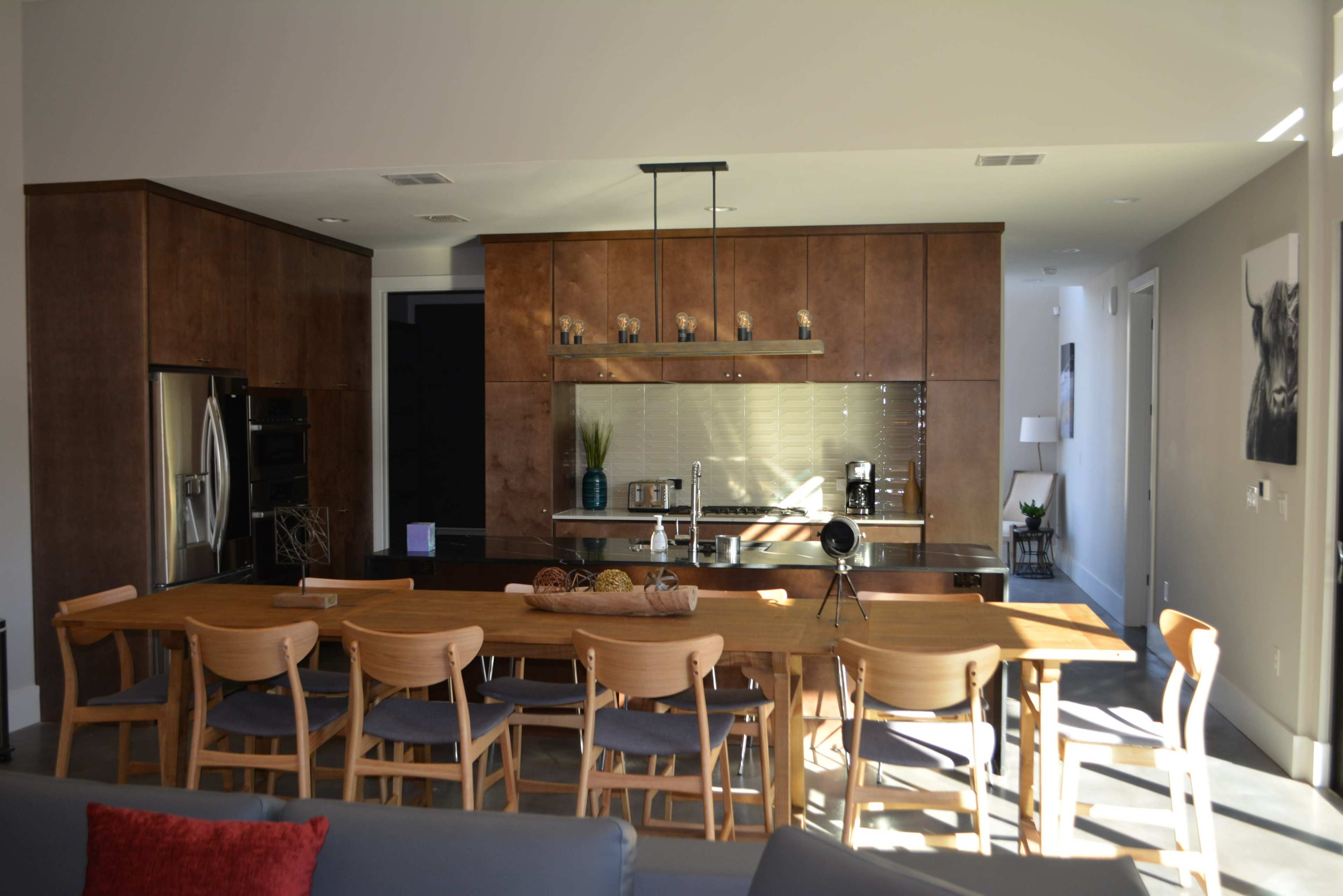 A modern kitchen features a long wooden dining table with eight chairs, surrounded by cabinets and a countertop with a minimalist design.