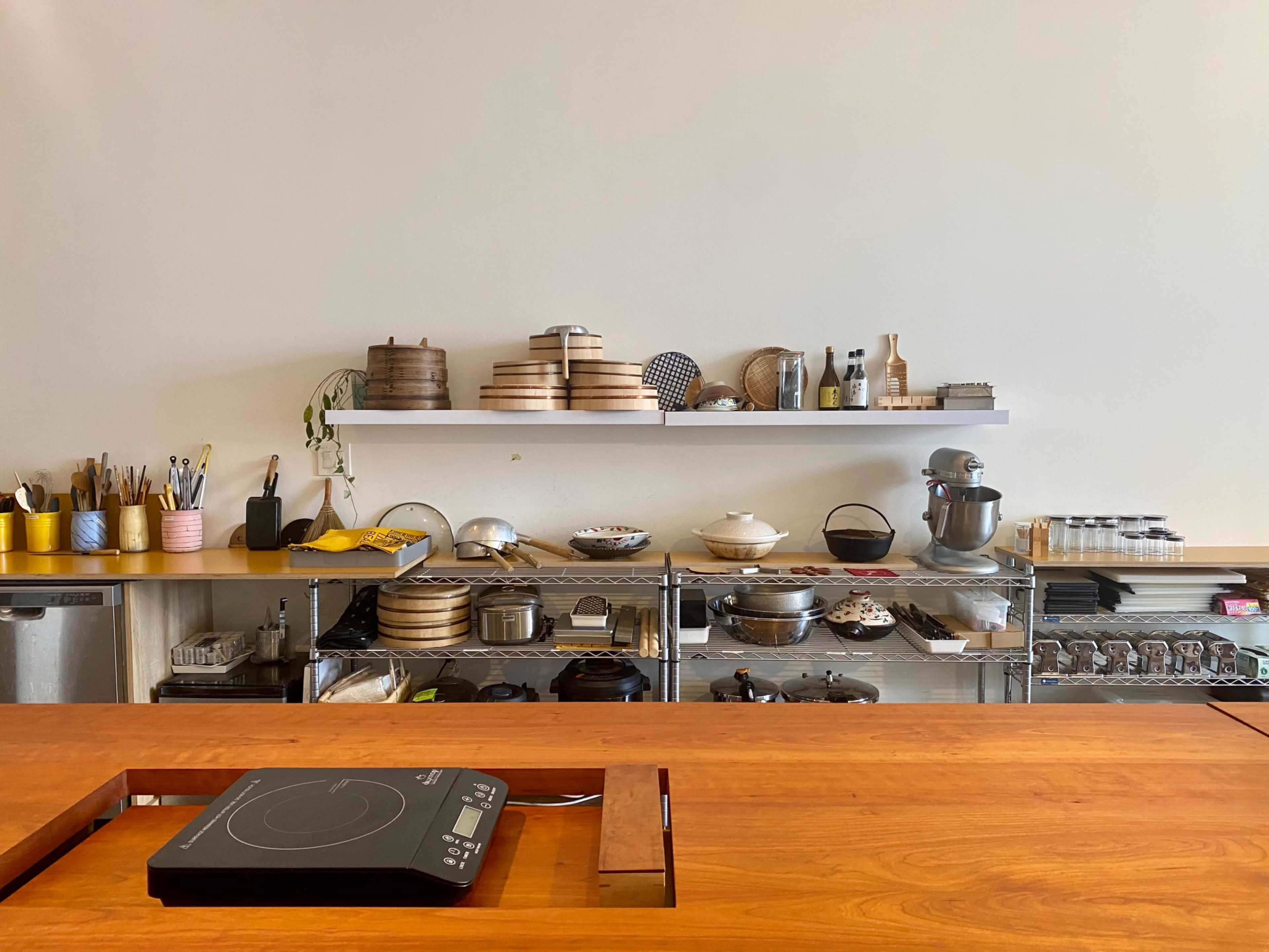 The image shows a kitchen with a wooden counter and shelves filled with various cooking utensils, pots, and containers.