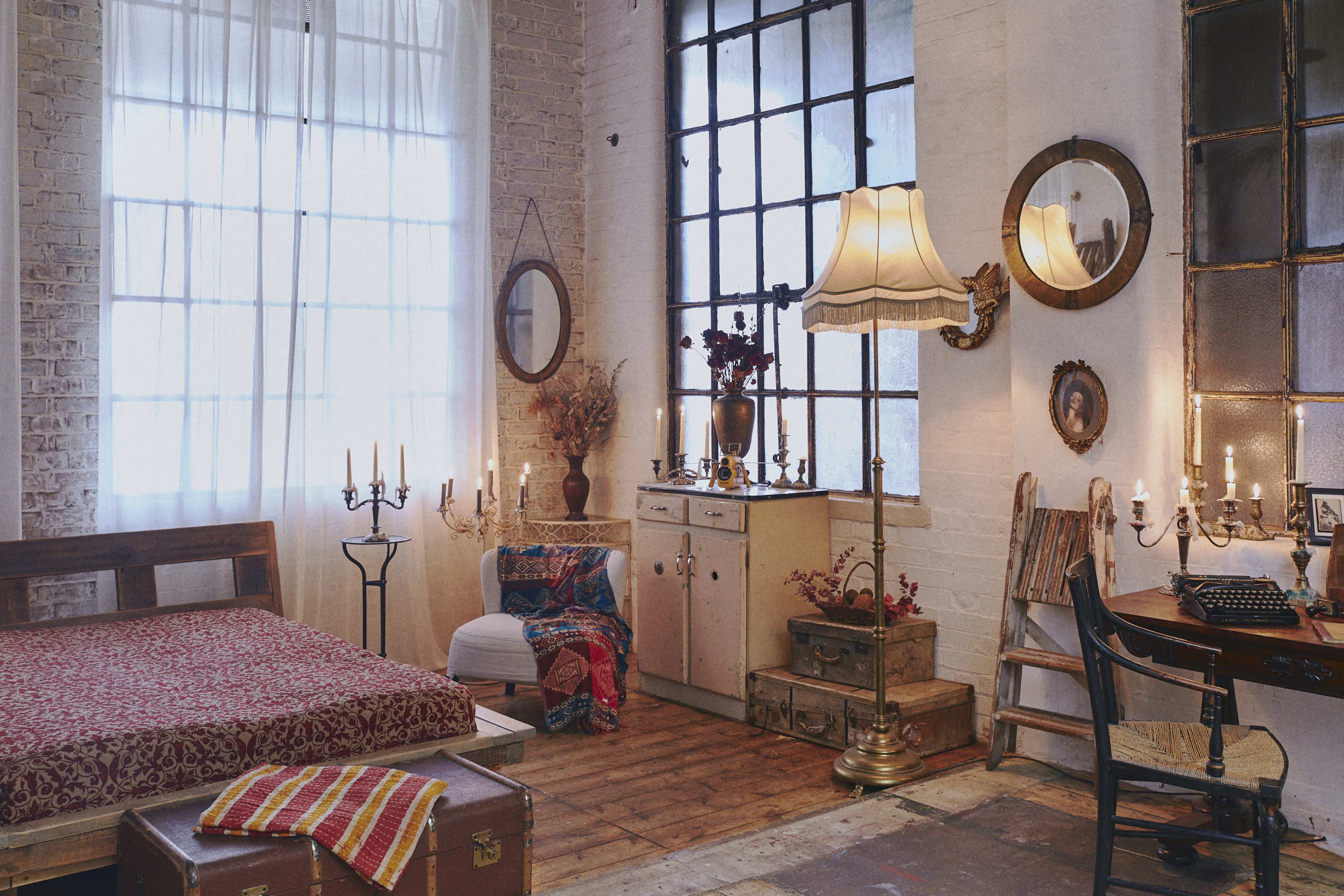 A cozy bedroom features a bed with a patterned cover, a wooden desk, vintage mirrors, and various decorative items against a backdrop of large windows.