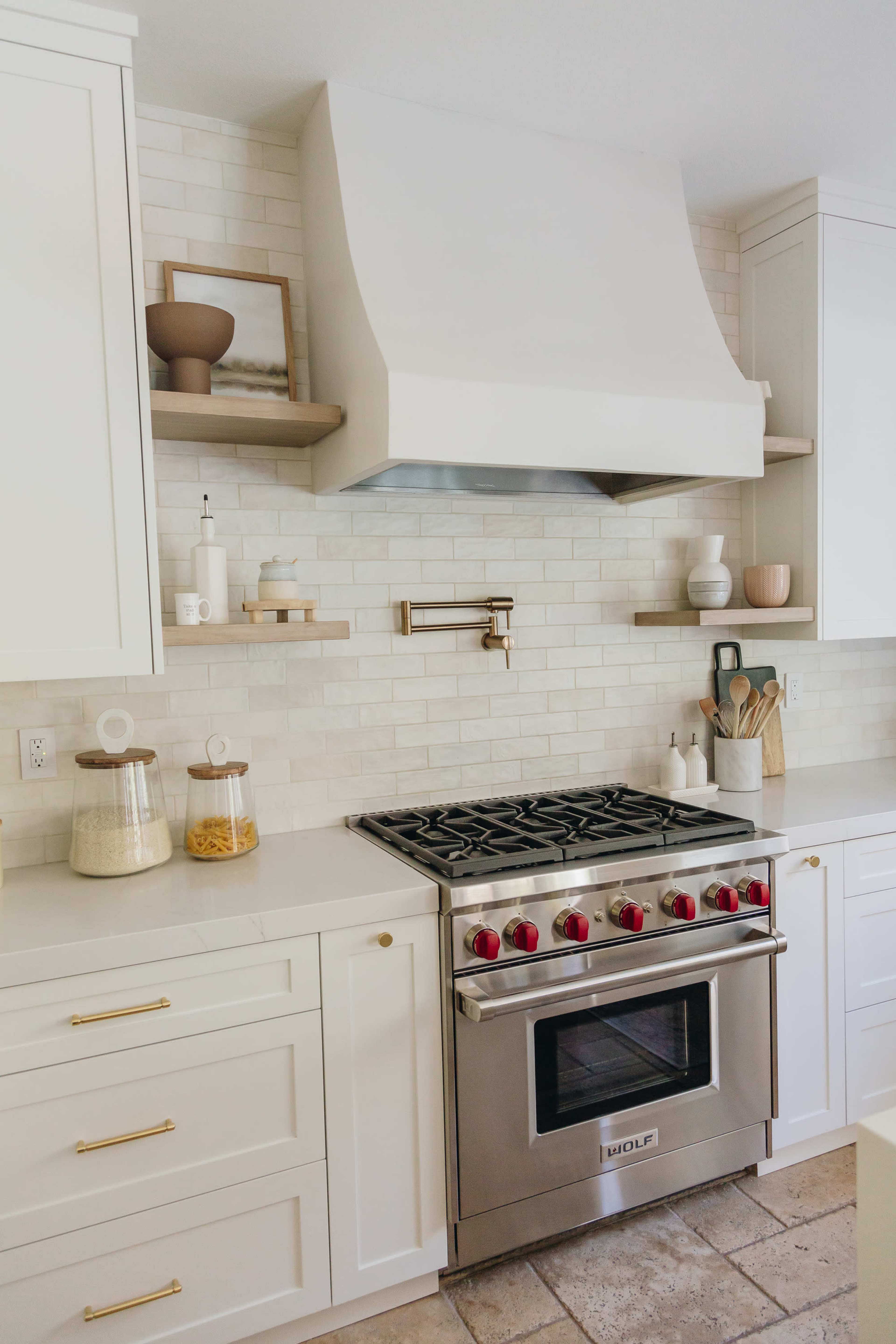 A modern kitchen features a stainless steel stove with red knobs below a white vent hood, surrounded by white cabinets and shelves holding various kitchen items.