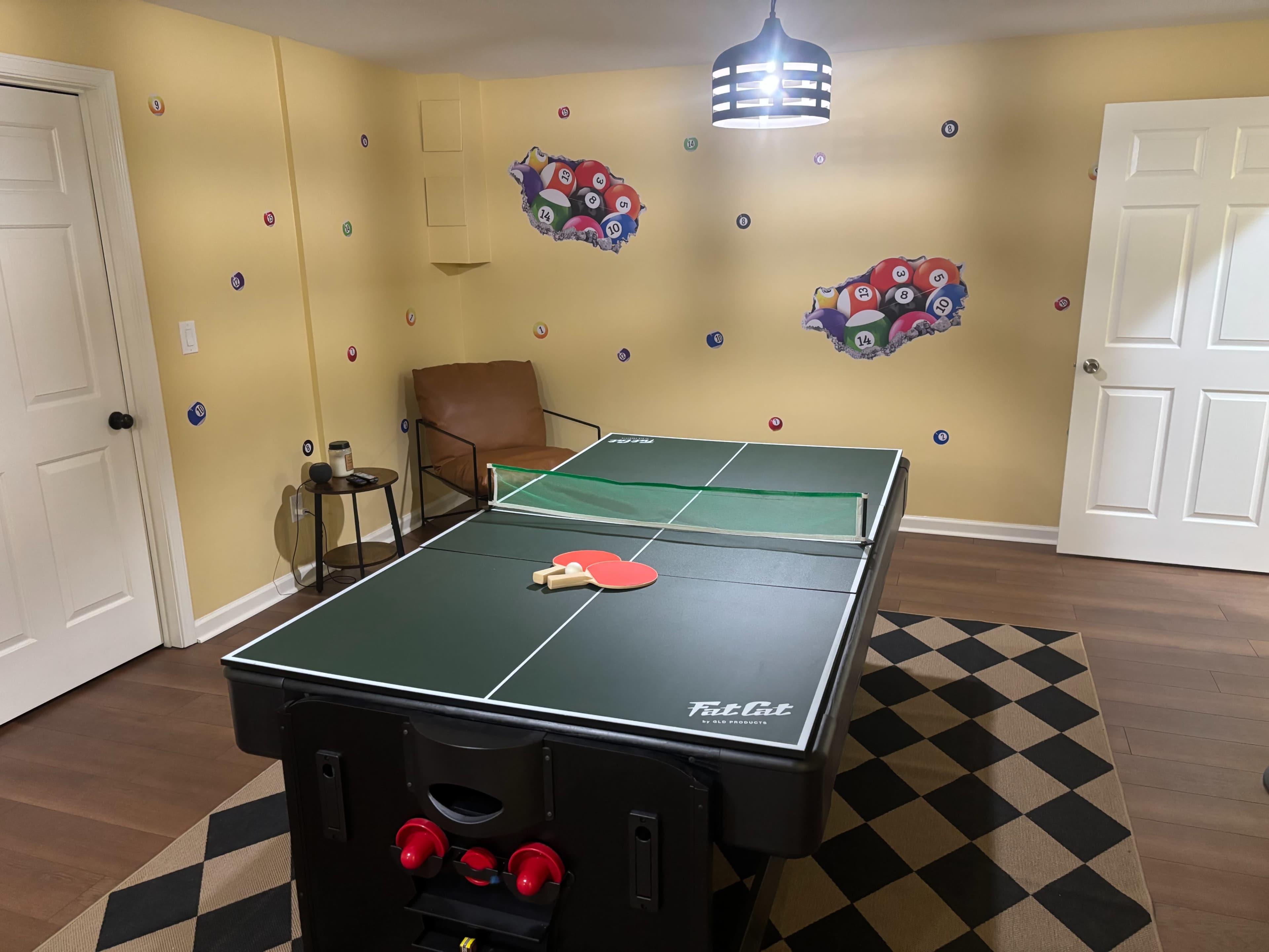 A ping pong table is set up in a room with yellow walls adorned with colorful wall decals, accompanied by a brown chair and a checkered rug.