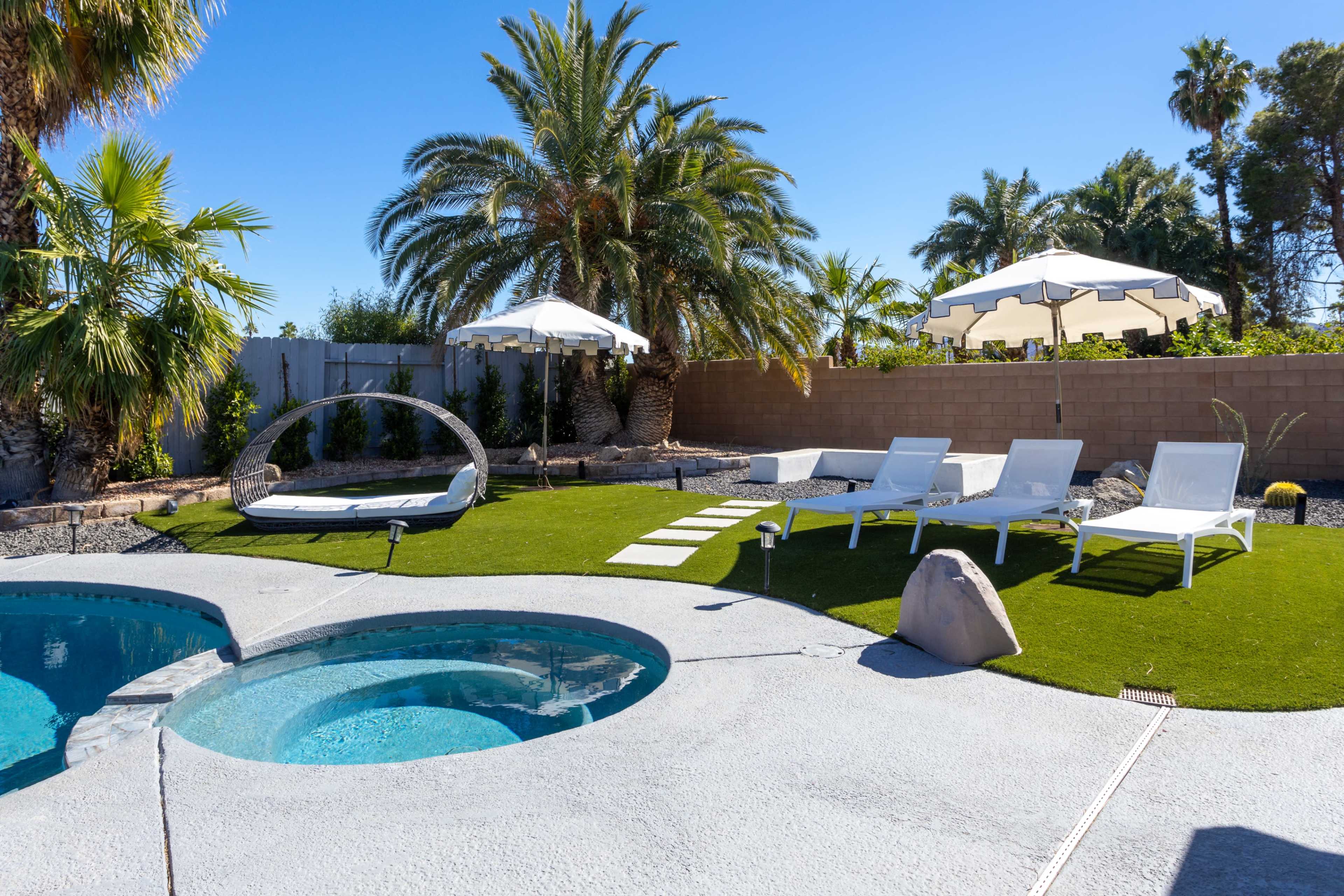 A backyard features a swimming pool with a hot tub, surrounded by lounge chairs, artificial grass, and palm trees under a clear blue sky.