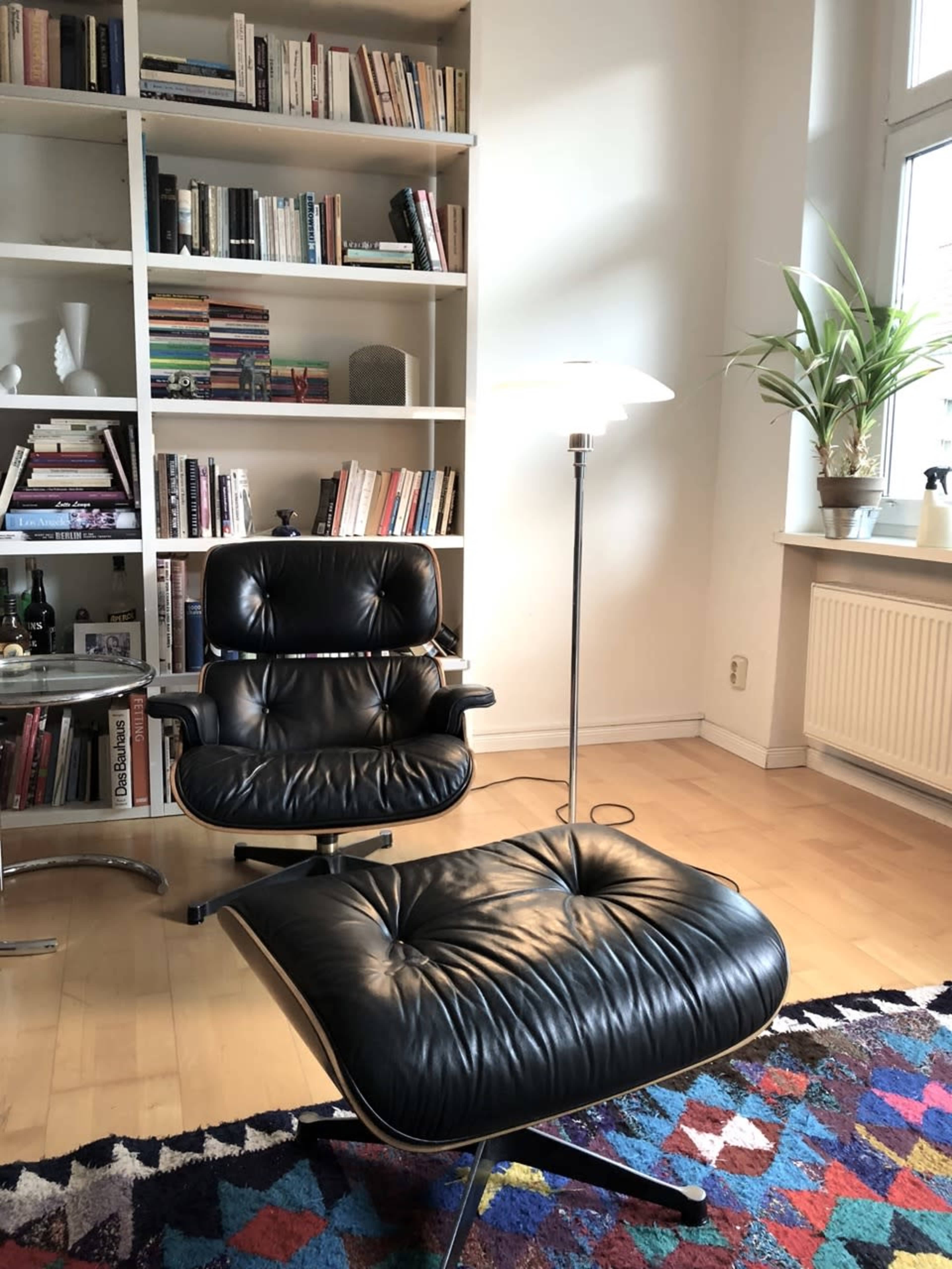 A black leather chair and ottoman sit next to a floor lamp in a room with a bookshelf filled with books and a potted plant.