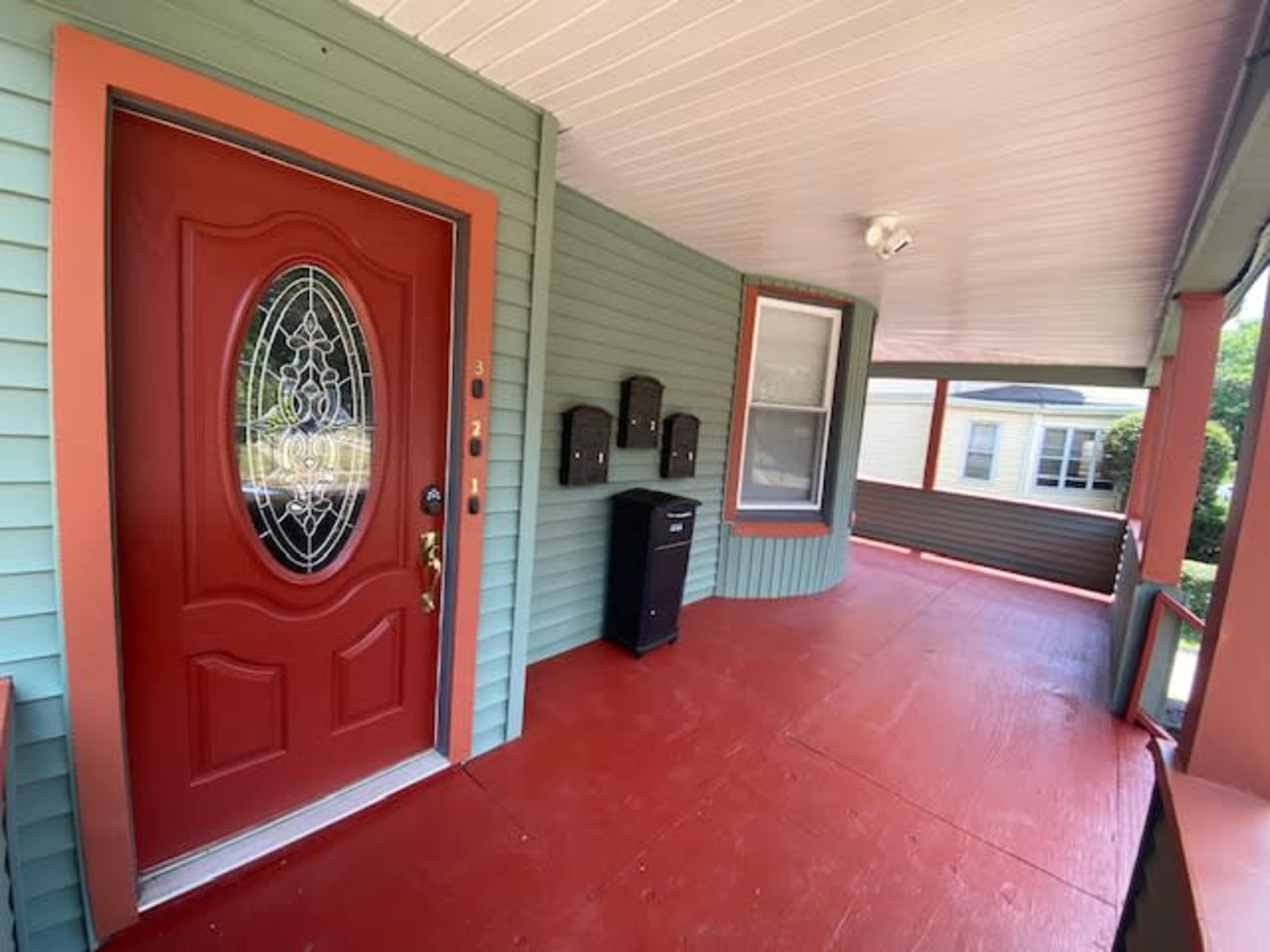 The image shows a porch with a red front door, green siding, and a black mailbox, featuring a wooden floor and several mailboxes mounted on the wall.