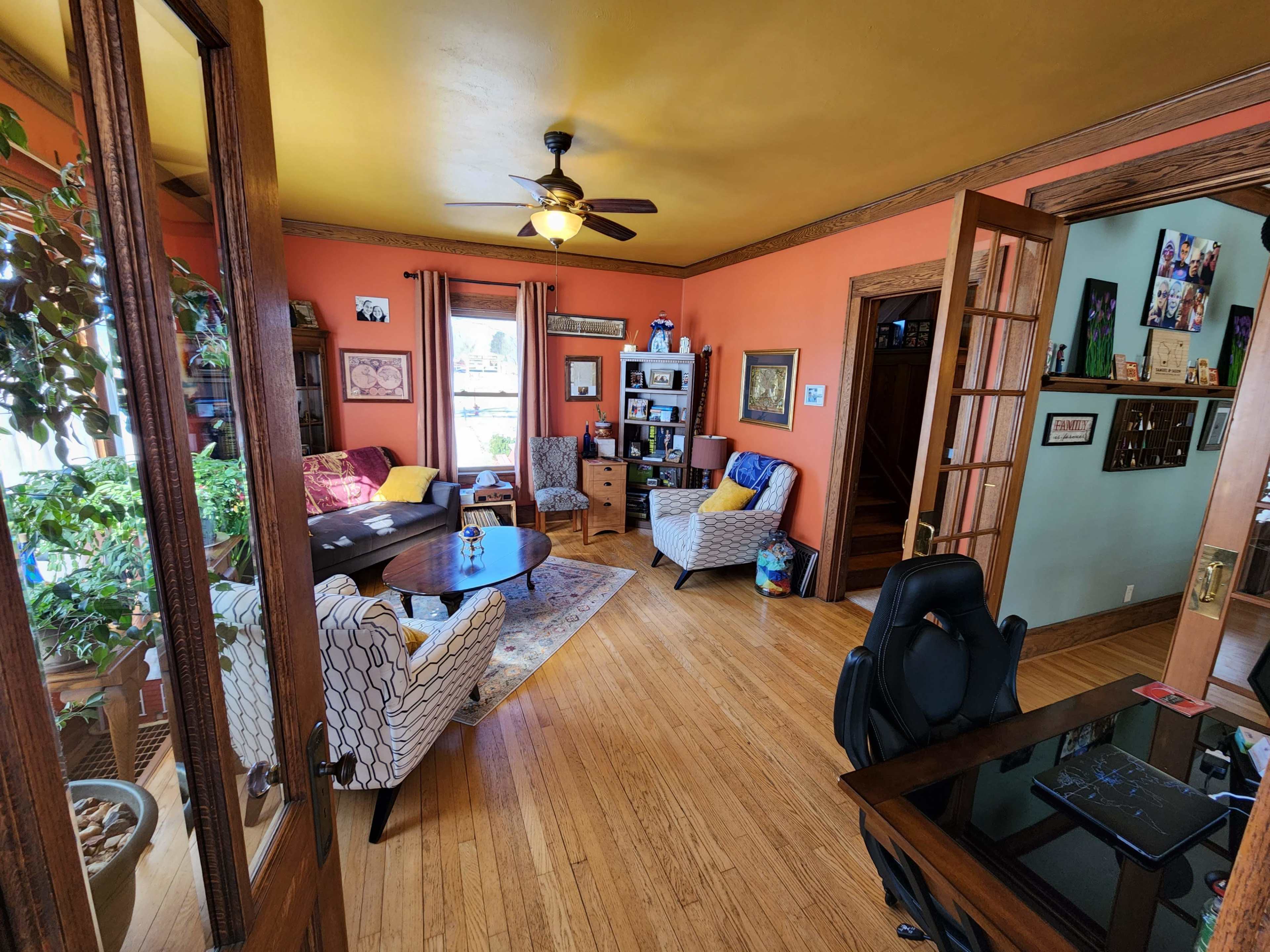 A living room with orange walls, hardwood floors, two patterned chairs, a coffee table, a potted plant, and a desk in the foreground.