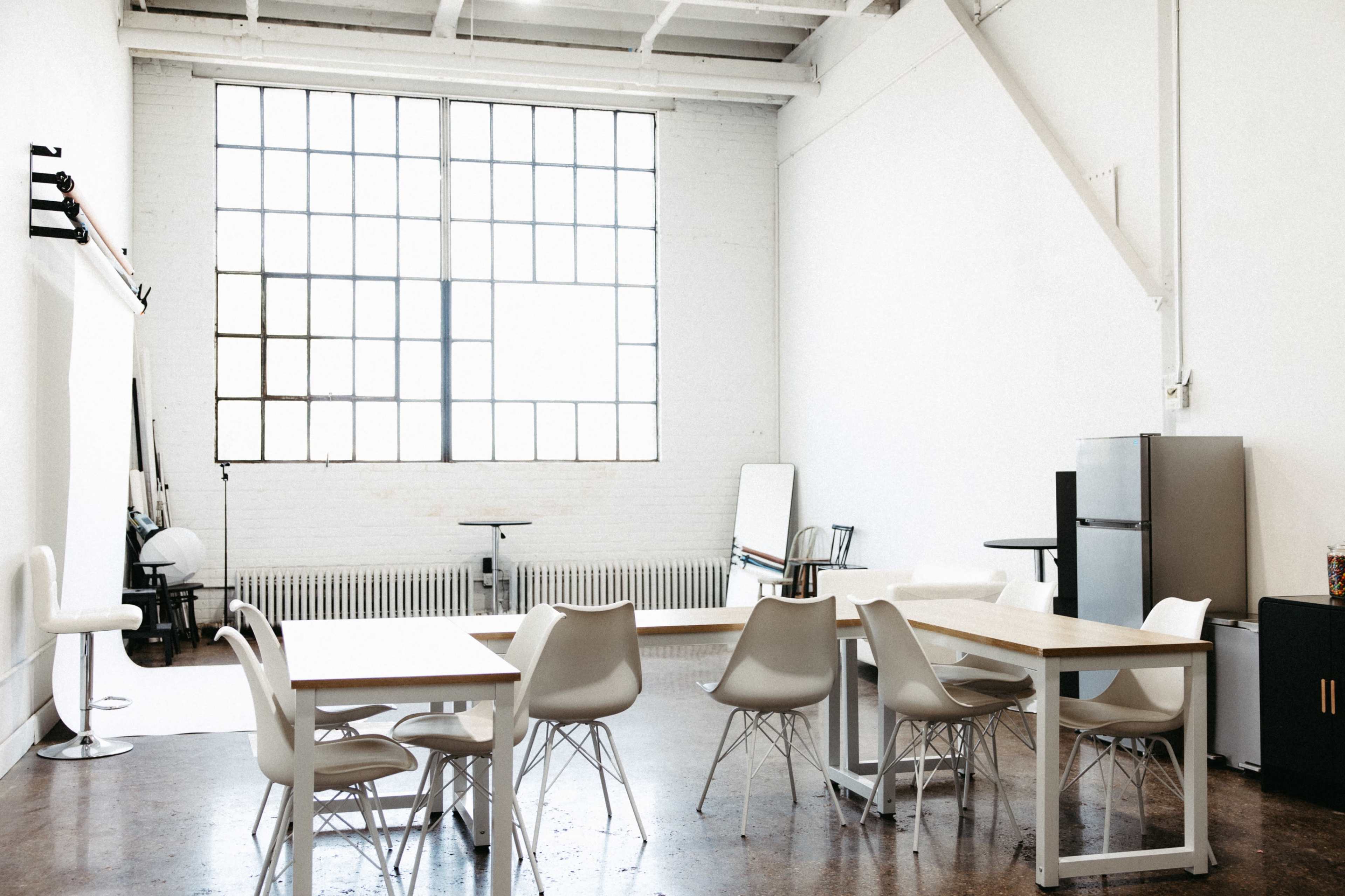 The image shows a spacious, minimalistic studio with large windows, several tables, and chairs, along with a kitchenette area.