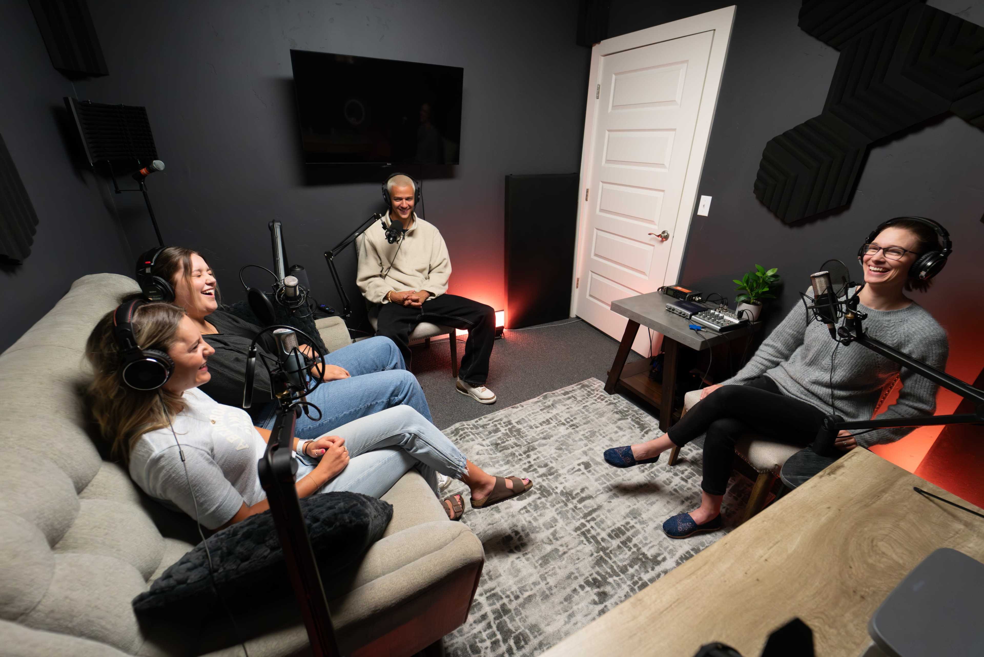A group of four individuals sits in a recording studio, engaged in conversation while using microphones and headphones.
