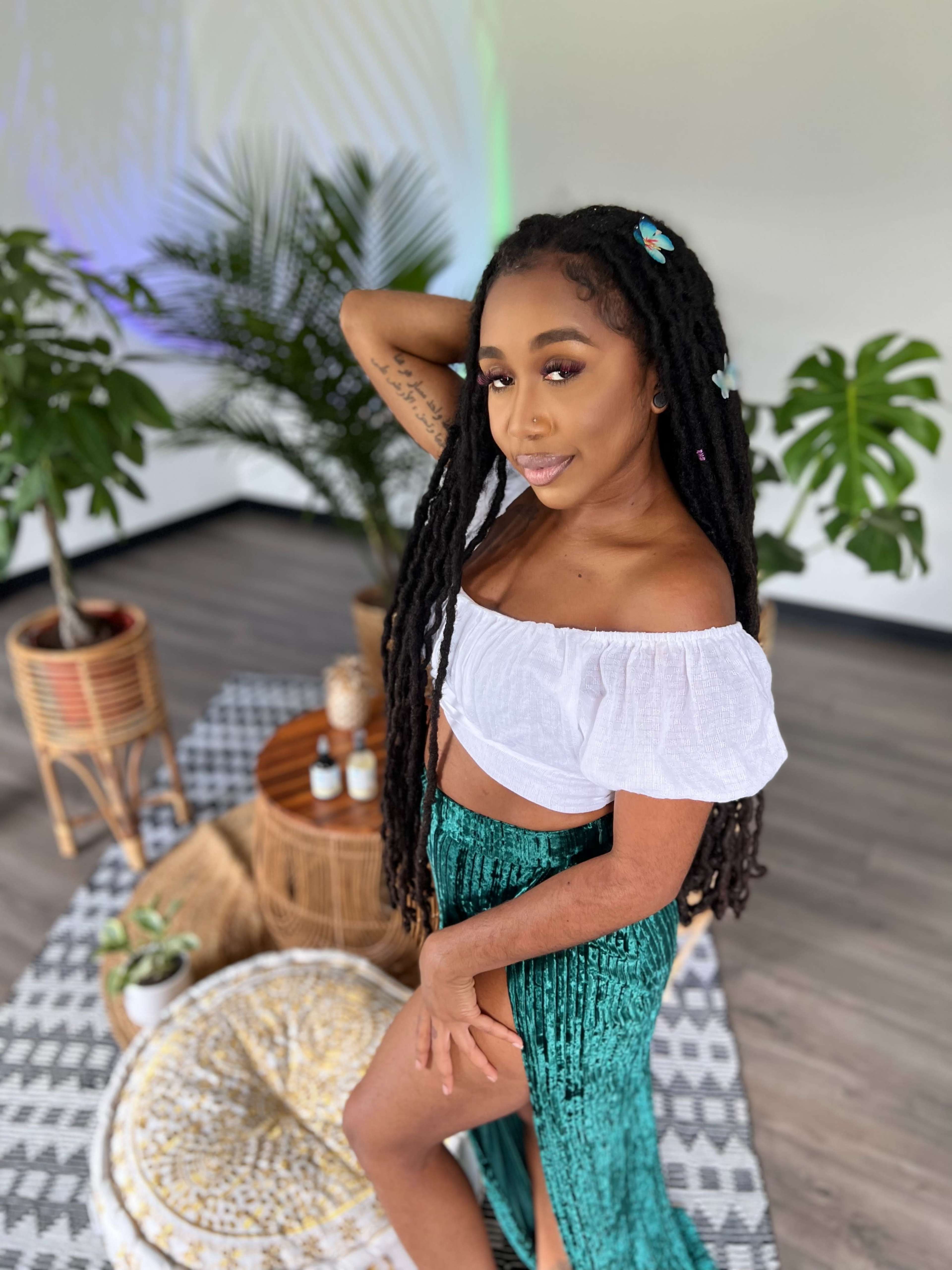 A woman with long black hair styled in braids poses by a green plant, wearing a white off-the-shoulder top and a shiny green skirt, in a room with patterned decor on the floor.