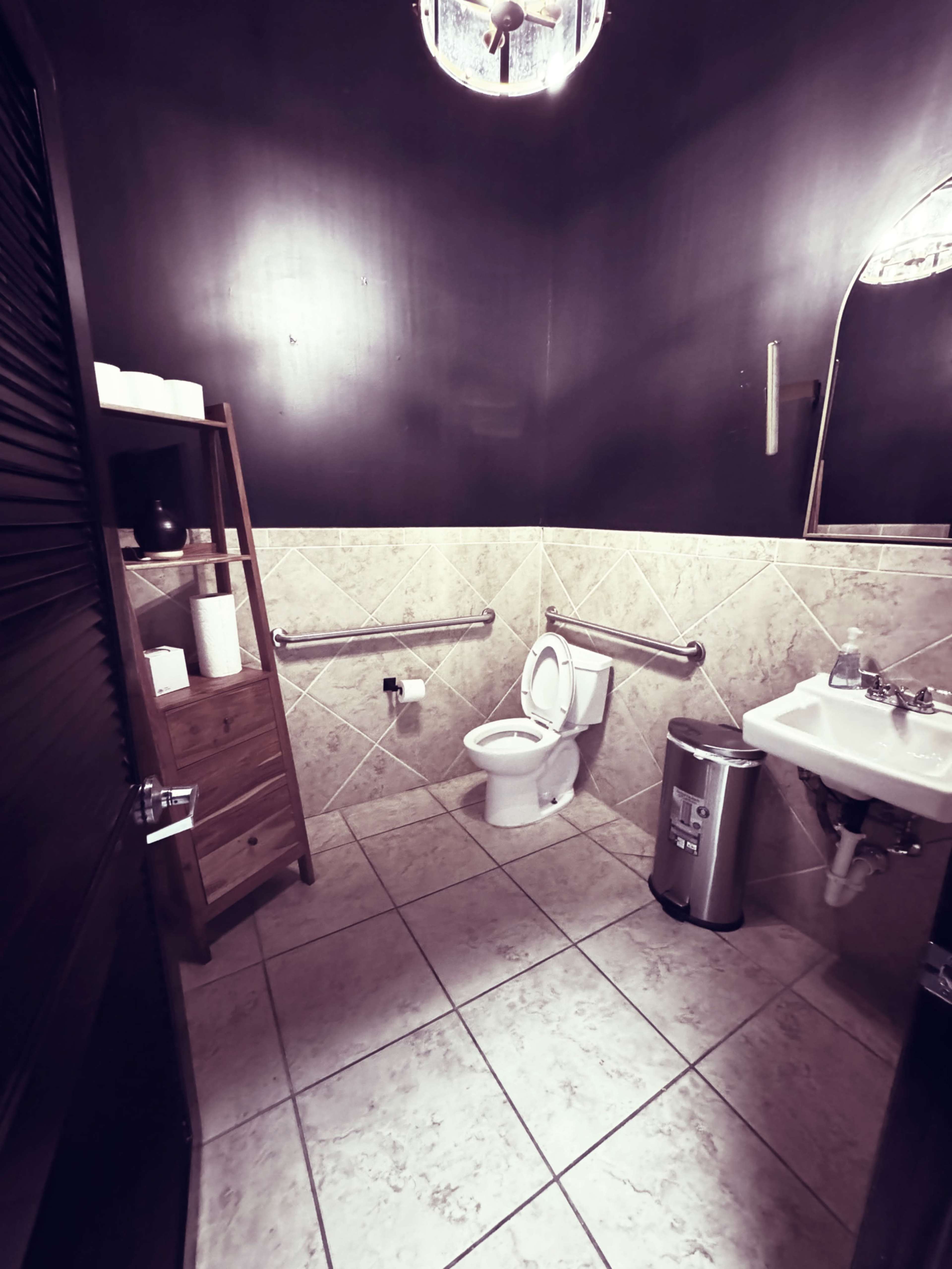 The image shows a small restroom with a toilet, sink, and a wooden storage unit, surrounded by dark walls and tiled flooring.