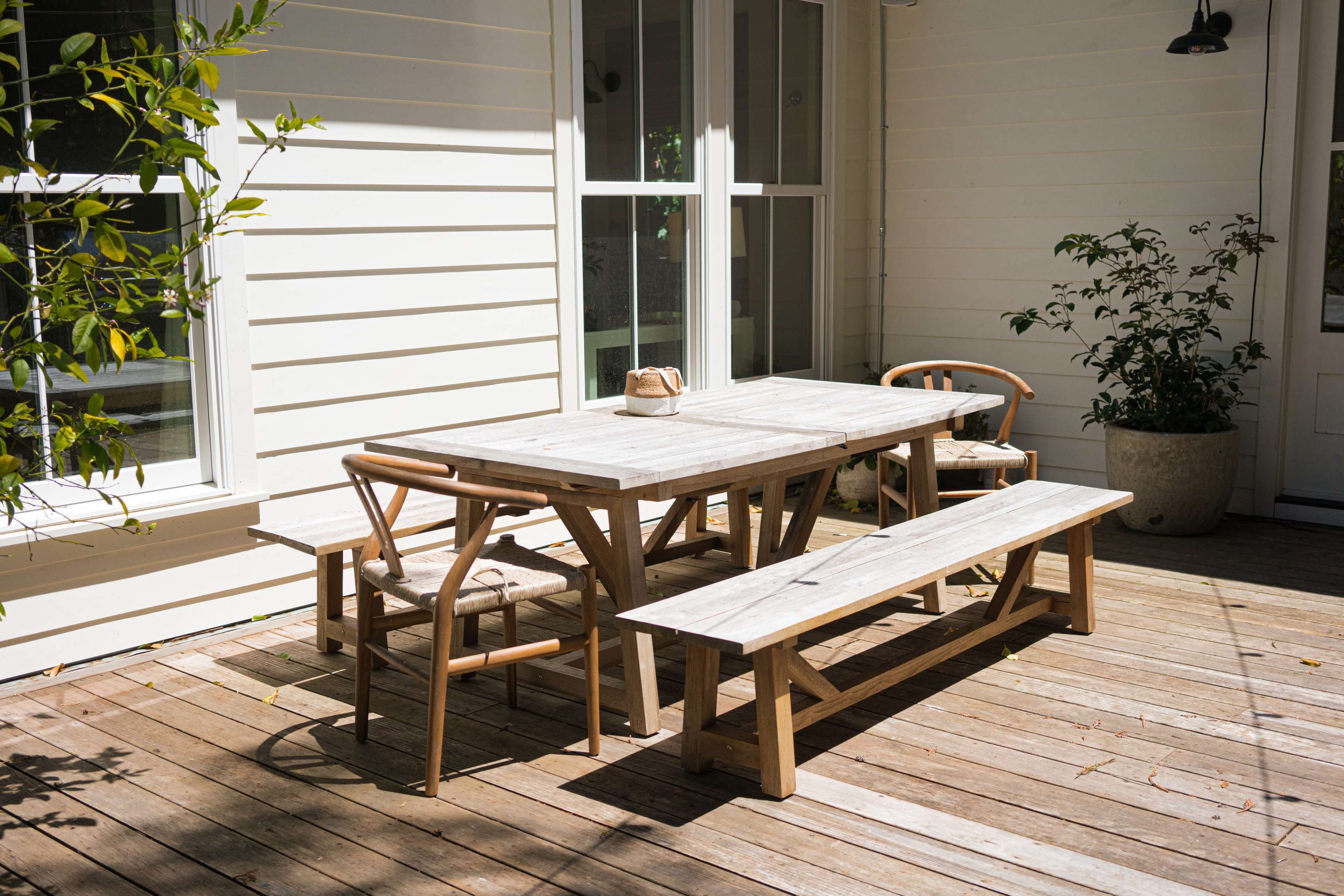 A wooden dining table surrounded by chairs and a bench is positioned on a wooden deck beside a house, with sunlight filtering through nearby plants.