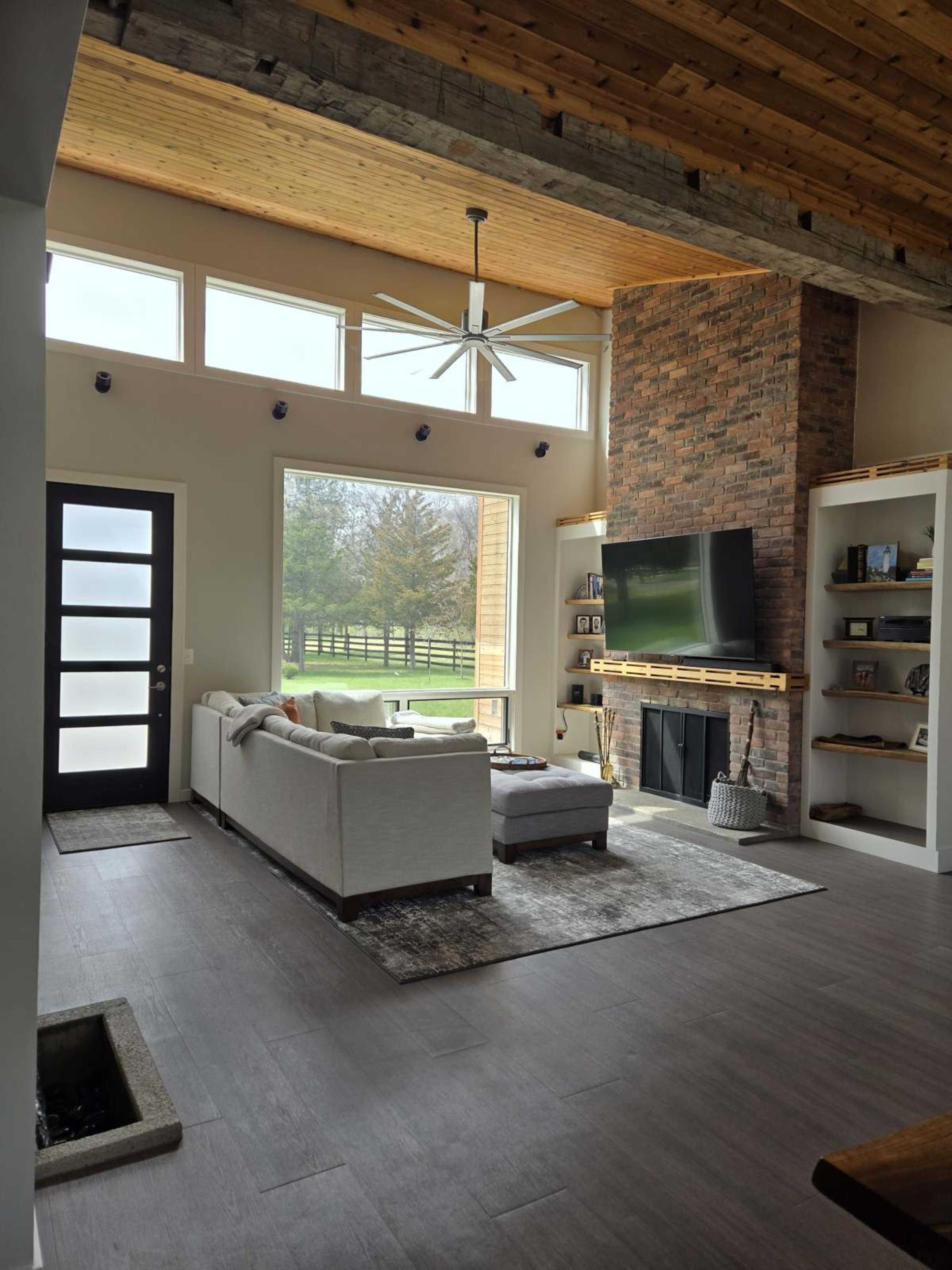 The image shows a modern living room with a large window, a comfortable sofa, a television mounted on a brick wall, and wooden ceiling beams.