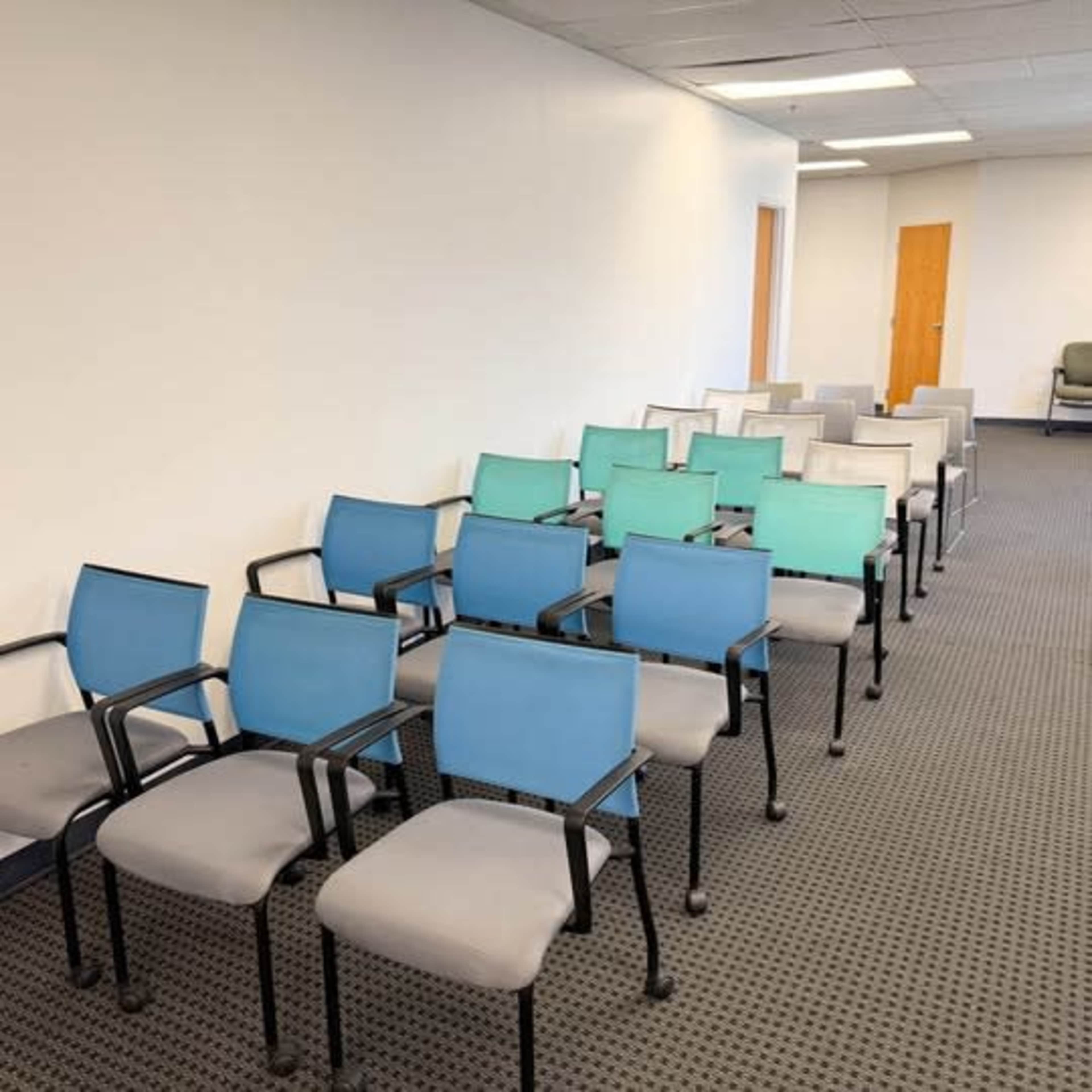 The image shows a row of multi-colored chairs set up in a sparse, well-lit room with beige walls and a carpeted floor.