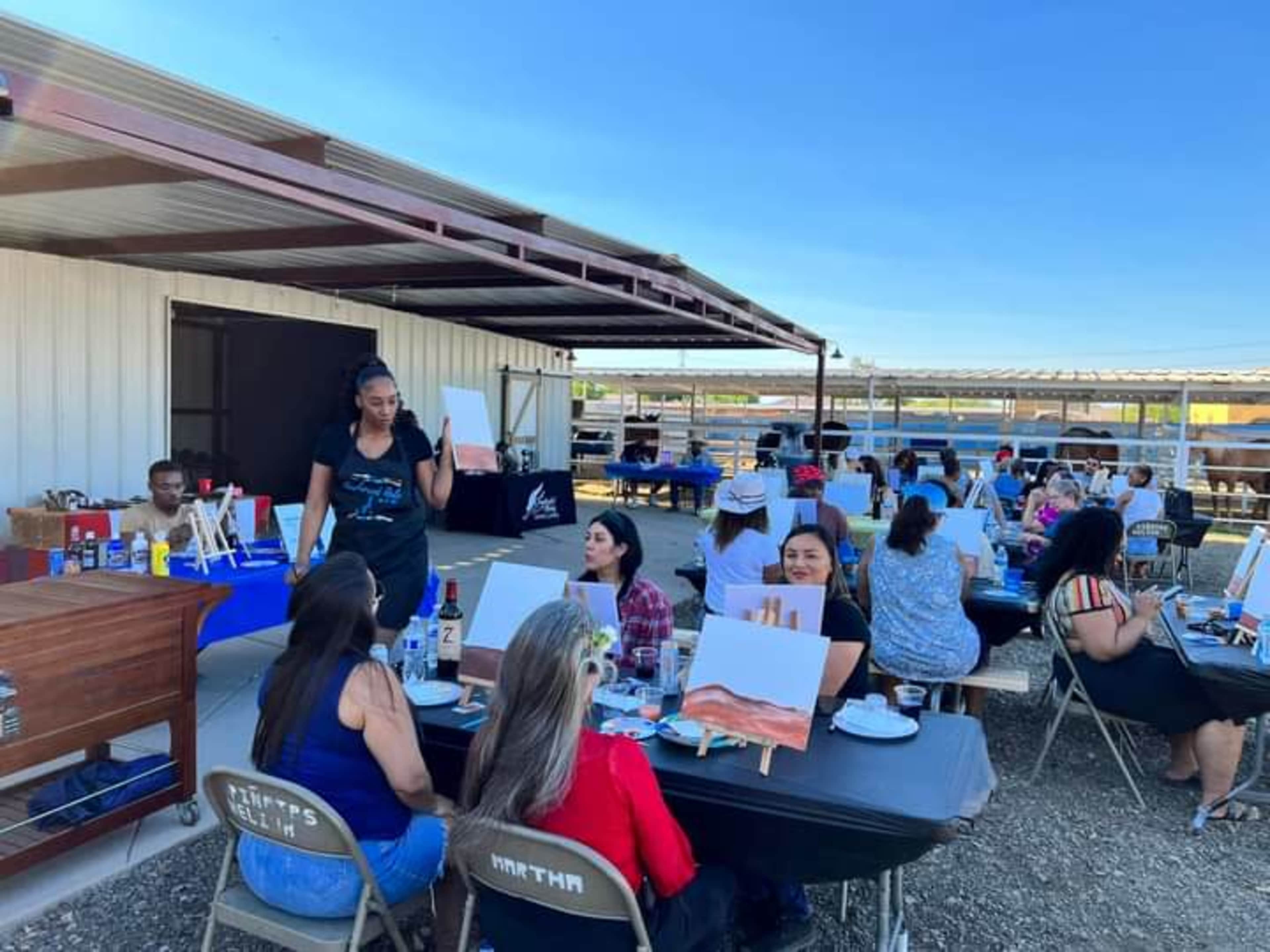 A group of people participate in a painting event outdoors, seated at tables with canvases and supplies under a covered area.