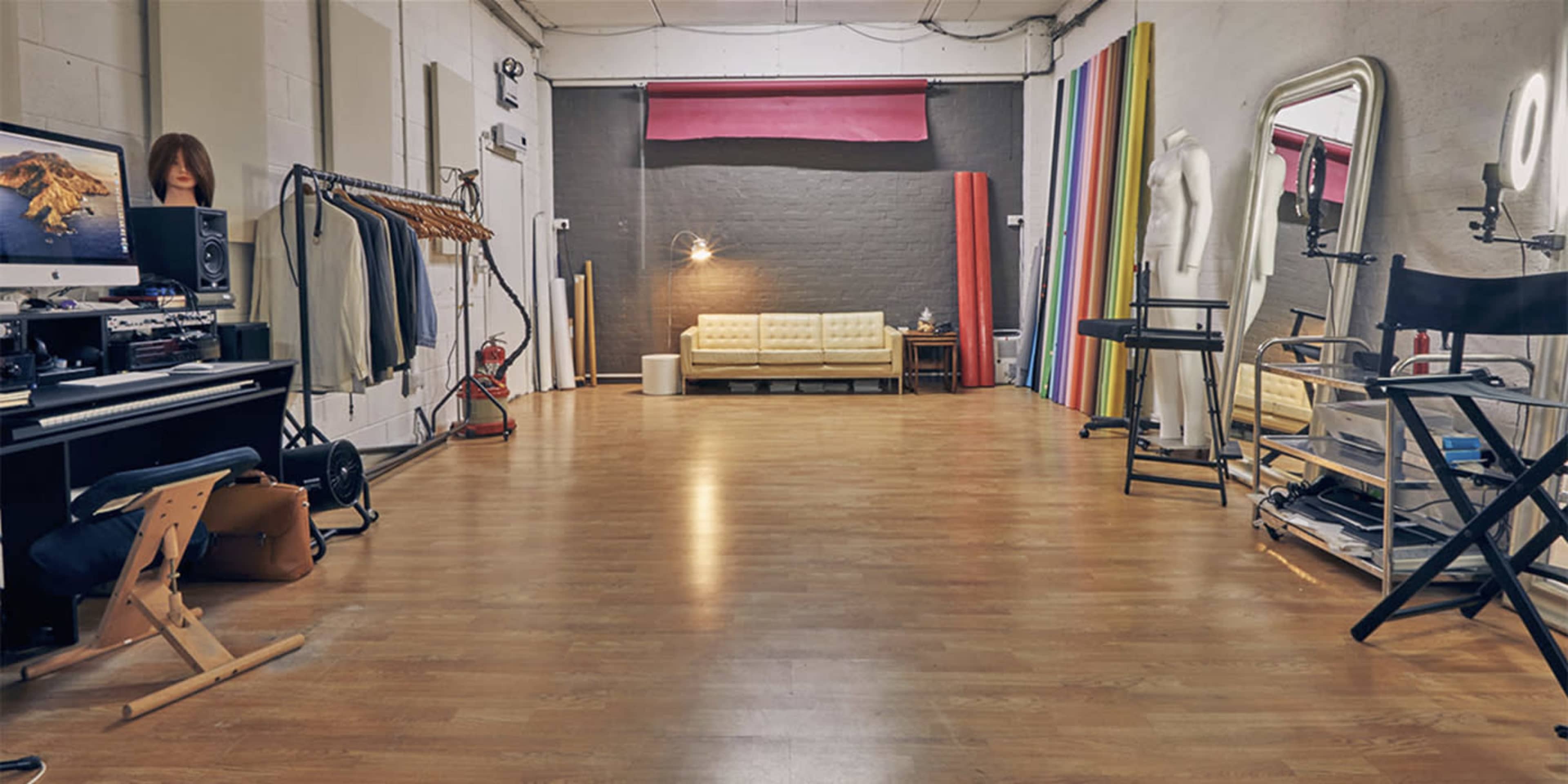 A spacious studio with wooden flooring, a seating area with a yellow couch, and various items including clothing racks, a mirror, and lighting equipment.