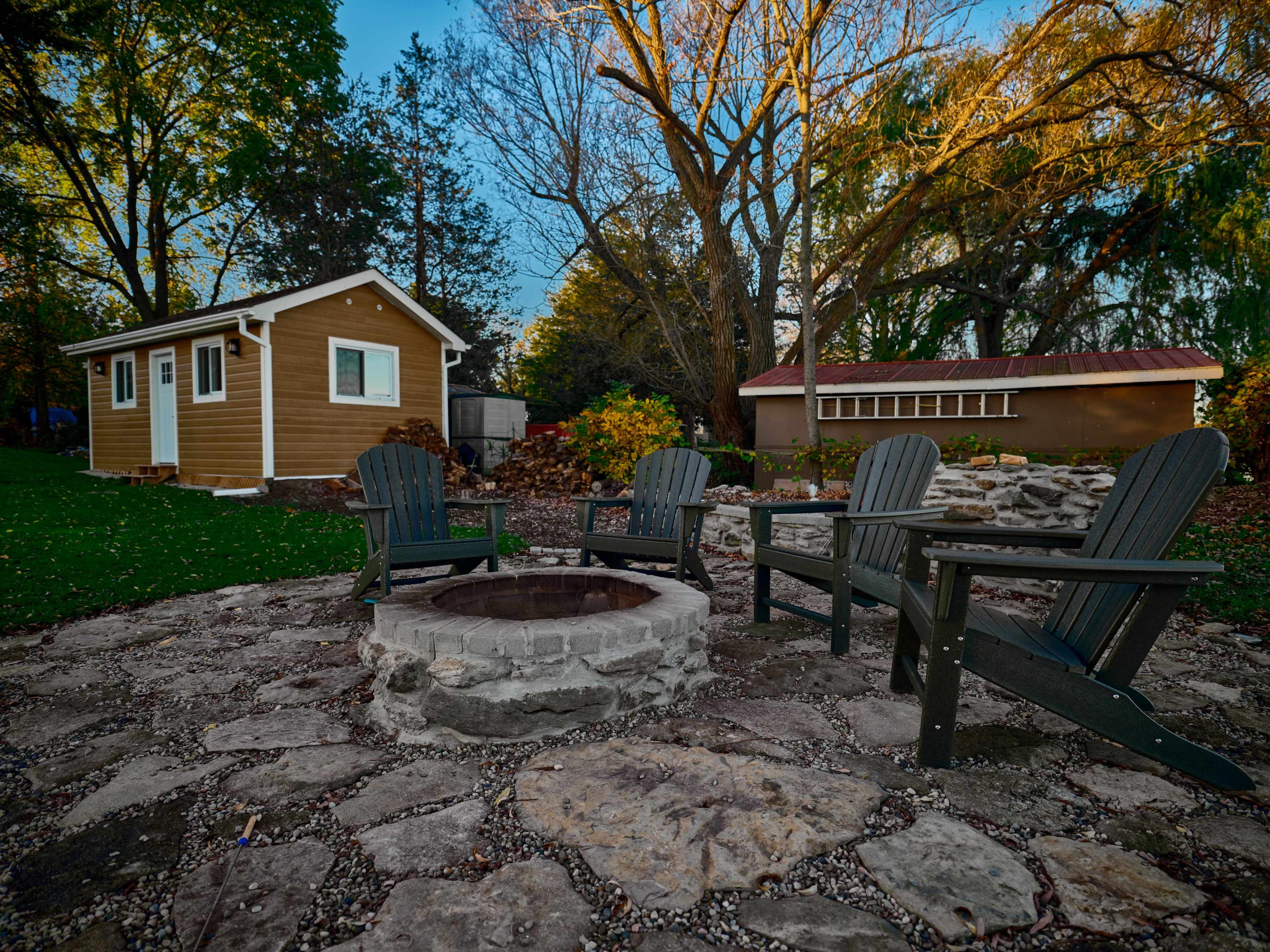 A stone fire pit surrounded by four wooden chairs in a yard with a small cabin and another building in the background.