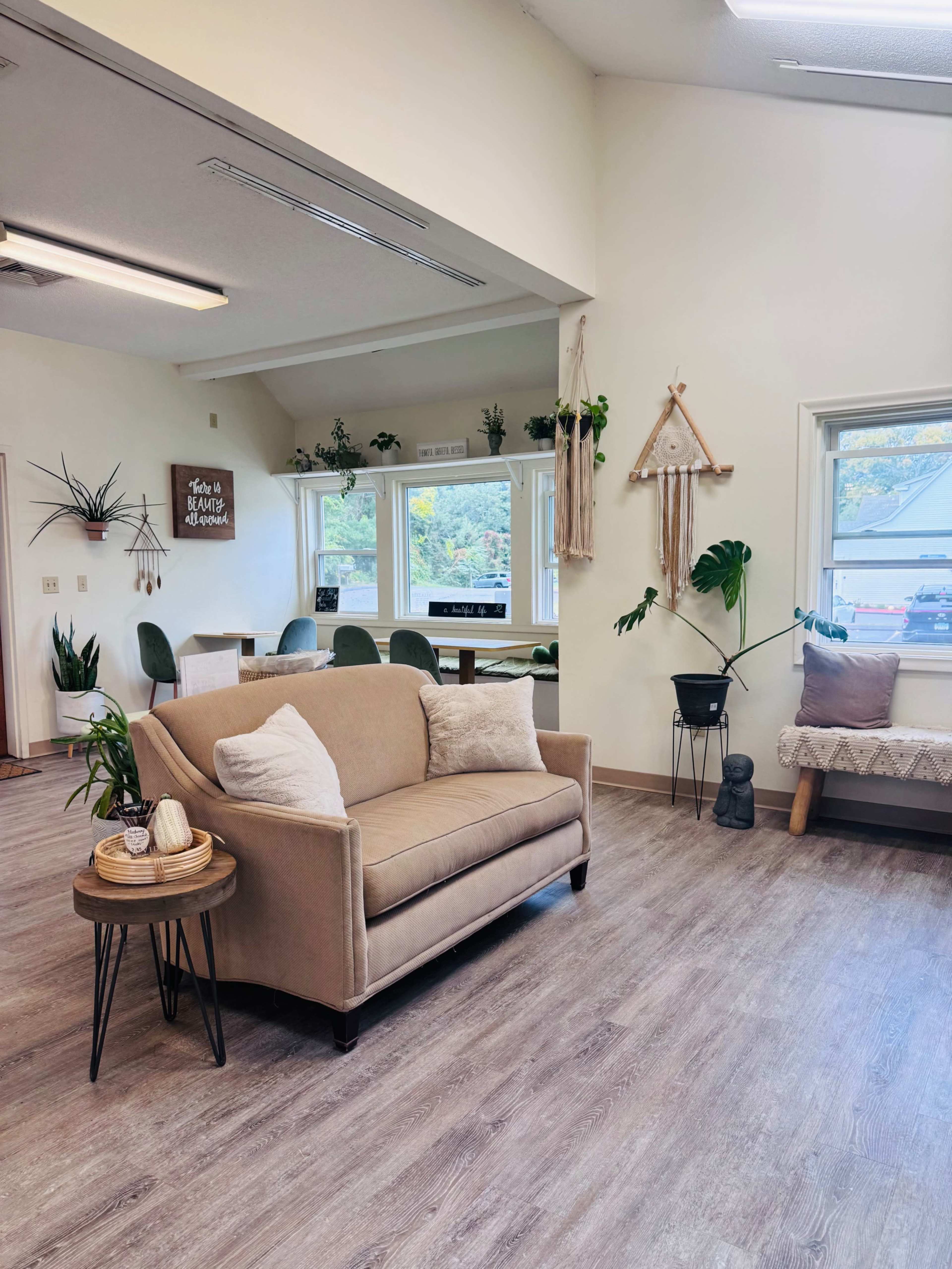 The image shows a cozy interior space featuring a beige couch, a small wooden table with decor, plants, and a dining area in the background.