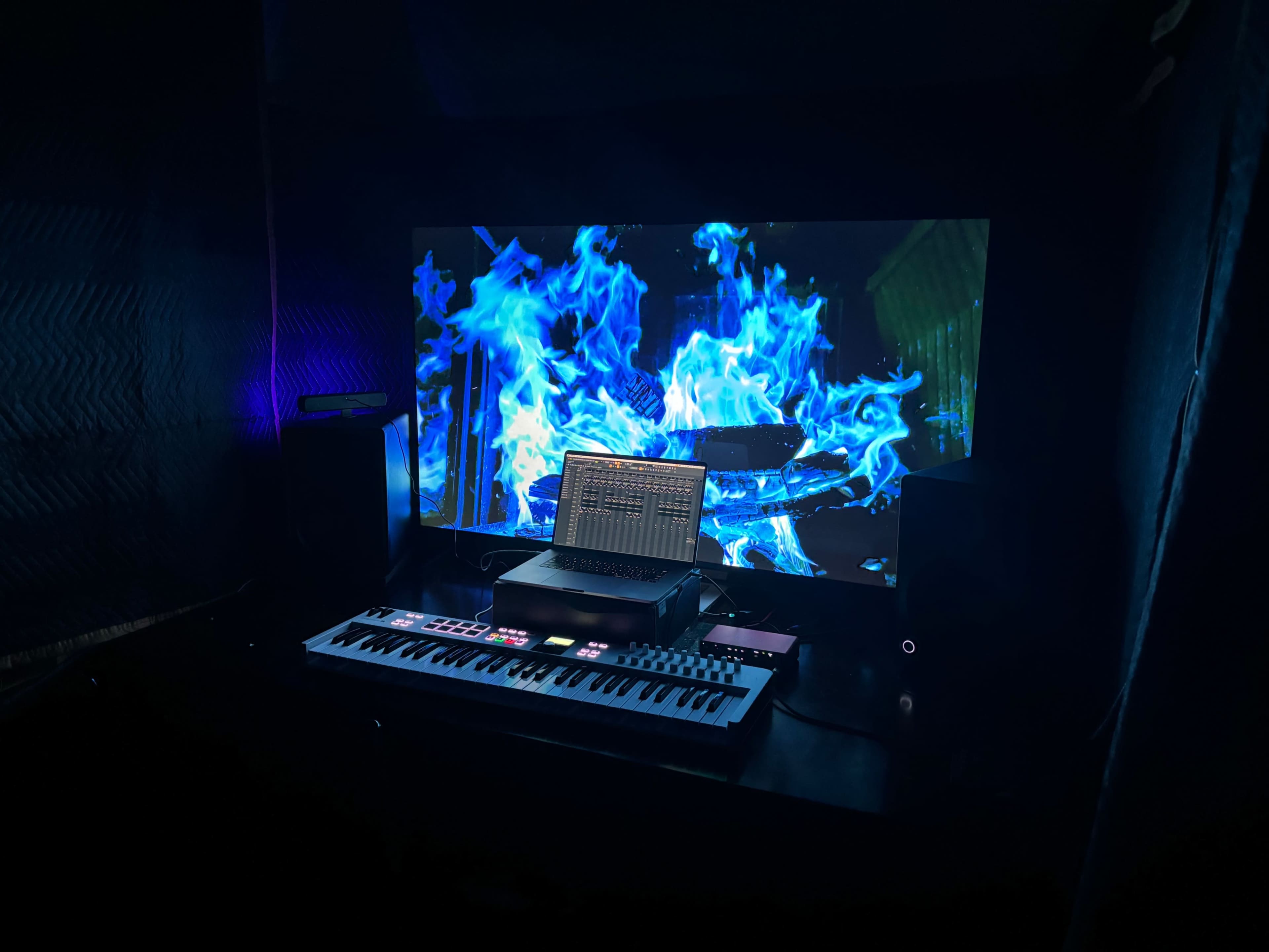 A digital music setup features a keyboard and laptop in front of a large screen displaying blue flames.