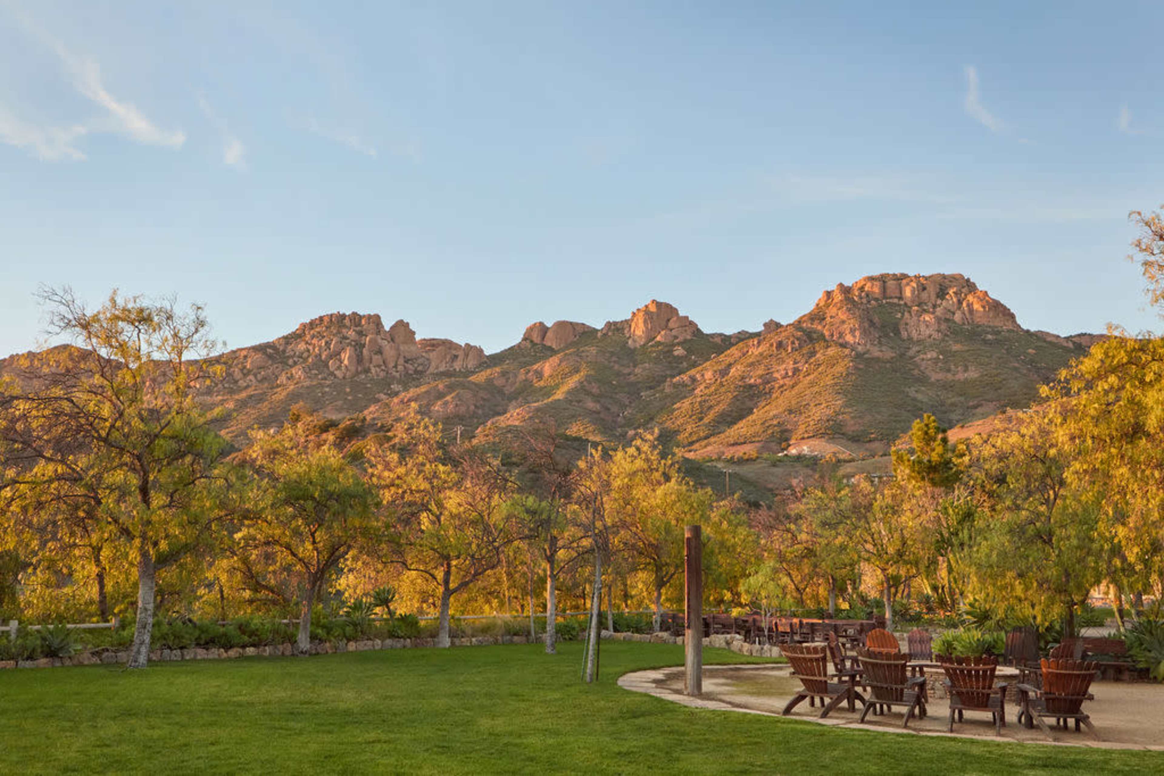A grassy area with wooden seating arrangements is set against a backdrop of rugged mountains under a clear sky.
