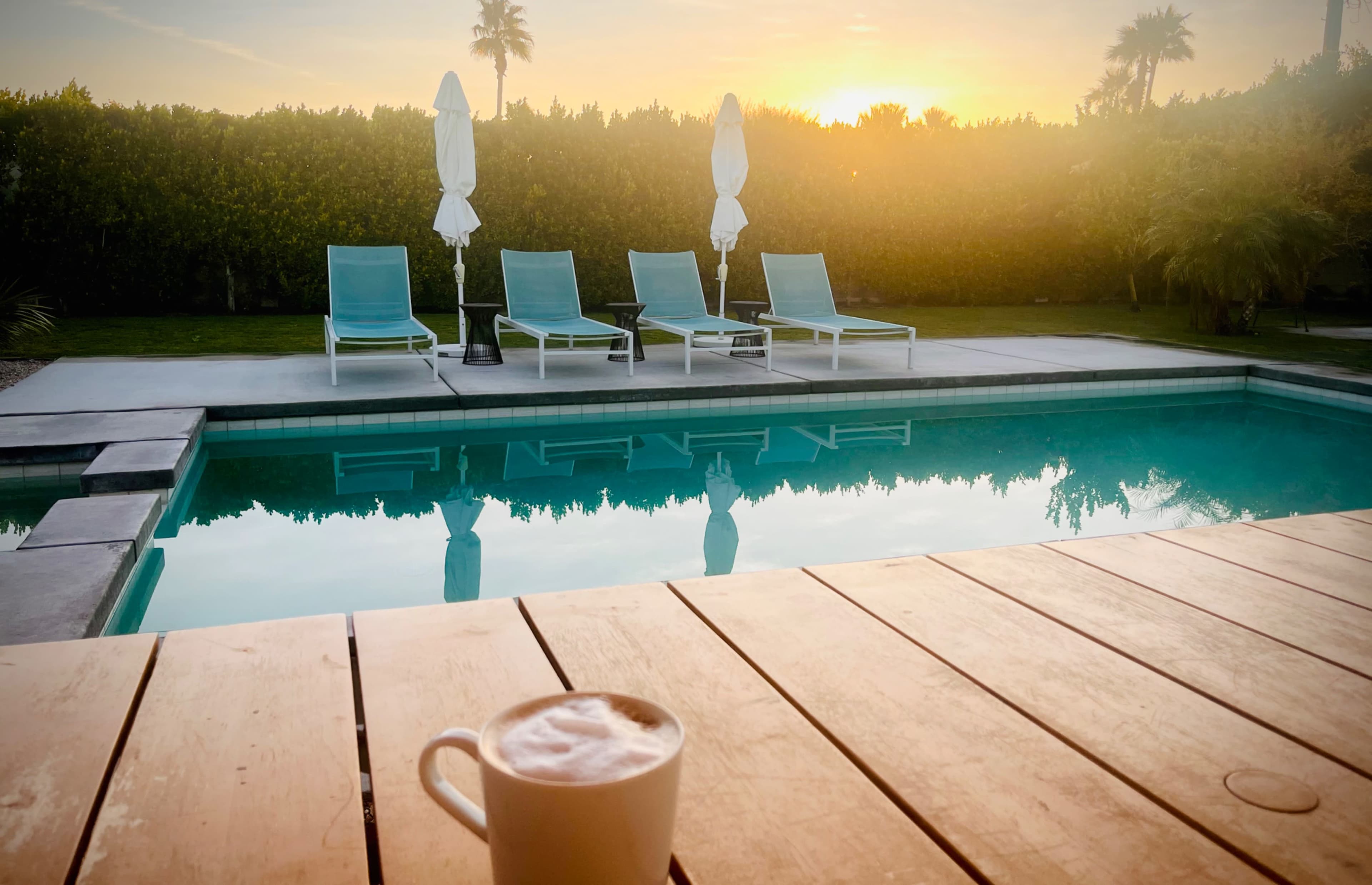 A mug of coffee sits on a wooden table in front of a swimming pool, with lounge chairs and palm trees visible in the background at sunset.