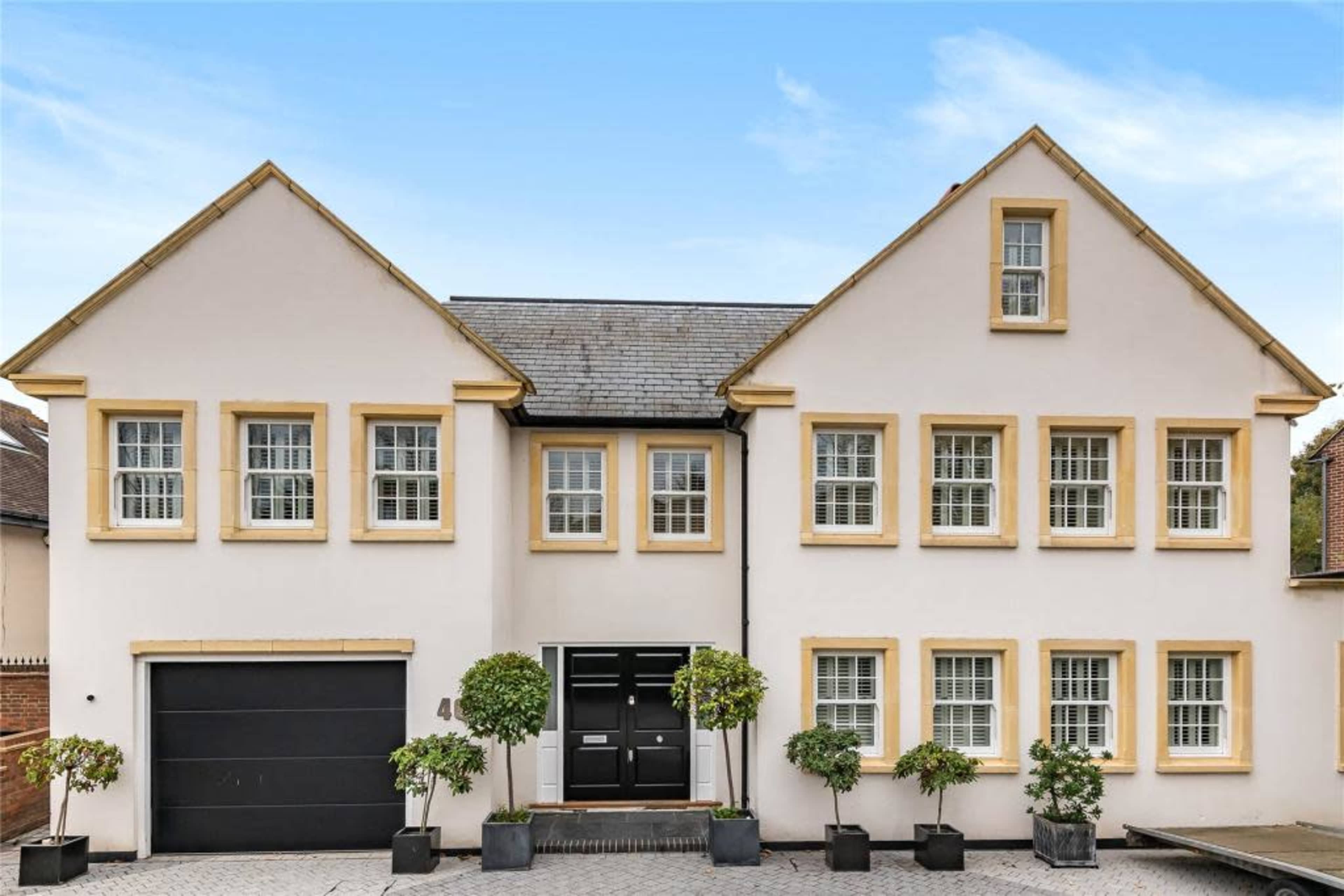 A two-story house features a symmetrical facade with six windows on the upper level and a central entrance, accompanied by neatly trimmed shrubs in planters flanking the door.