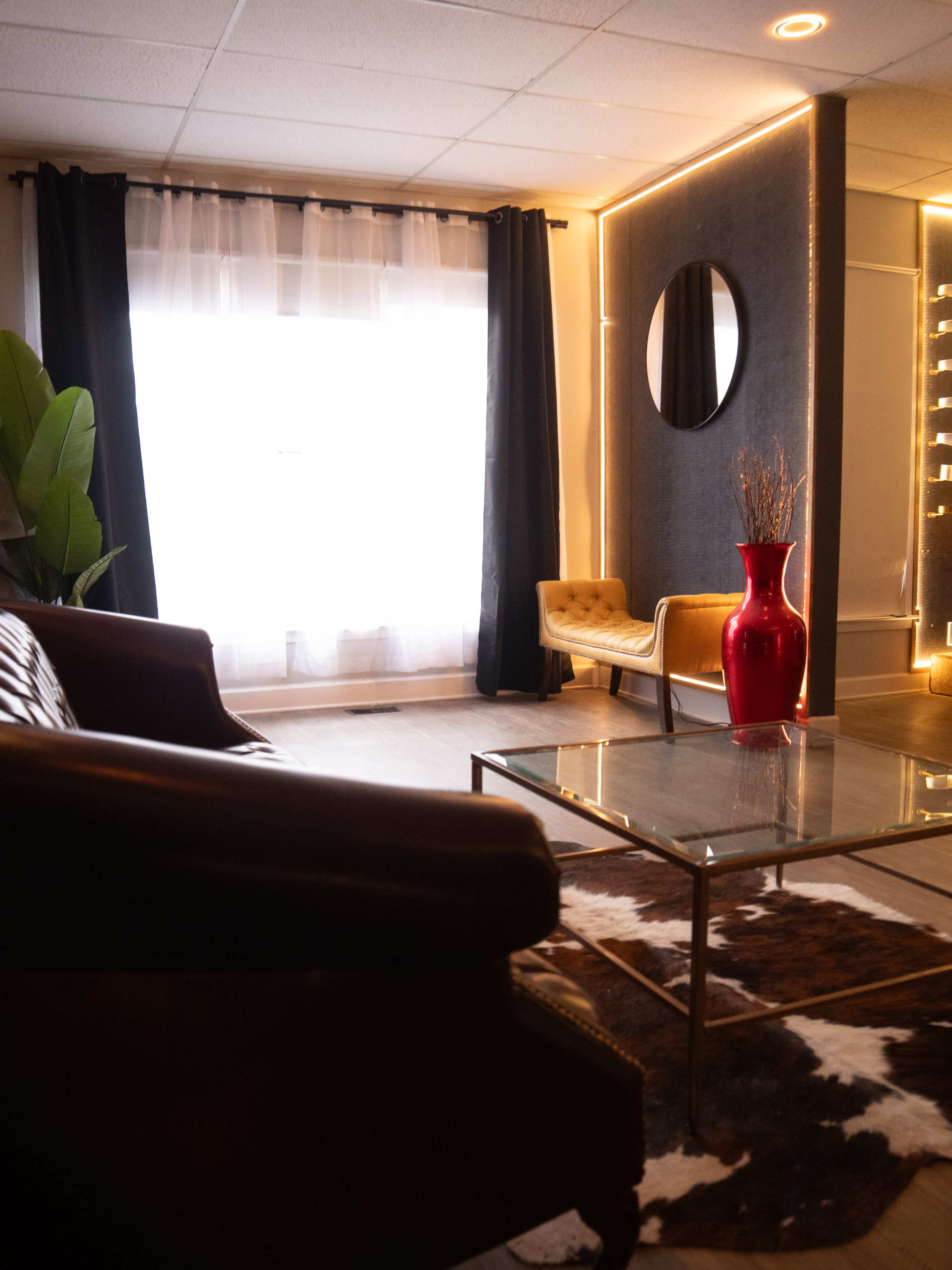 The image shows a cozy interior space featuring a leather sofa, a glass coffee table, a yellow armchair, a red vase, and large windows with sheer curtains.