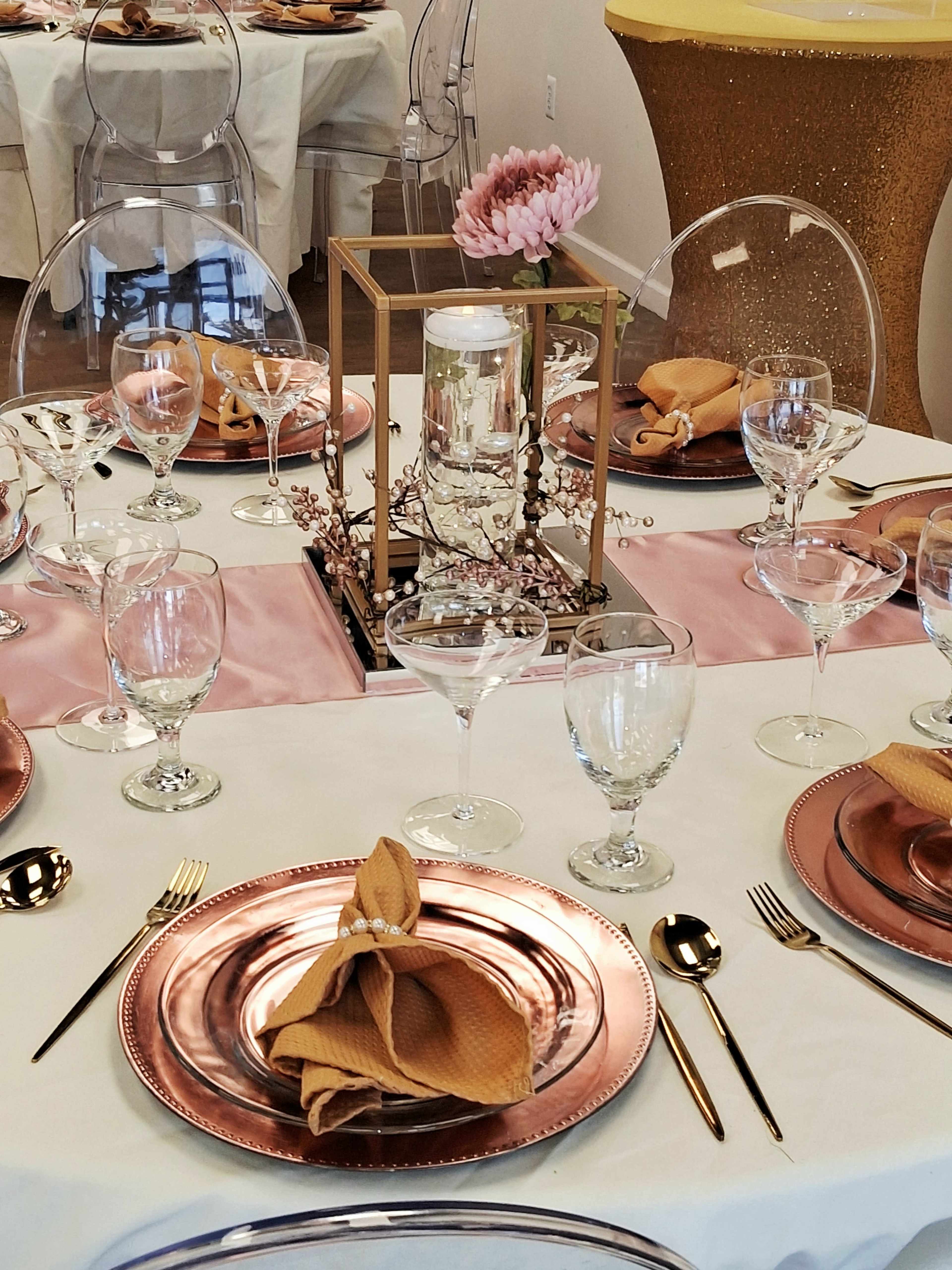 A beautifully set dining table features rose gold plates, elegant glassware, and a floral centerpiece, all arranged on a white tablecloth with soft pink accents.