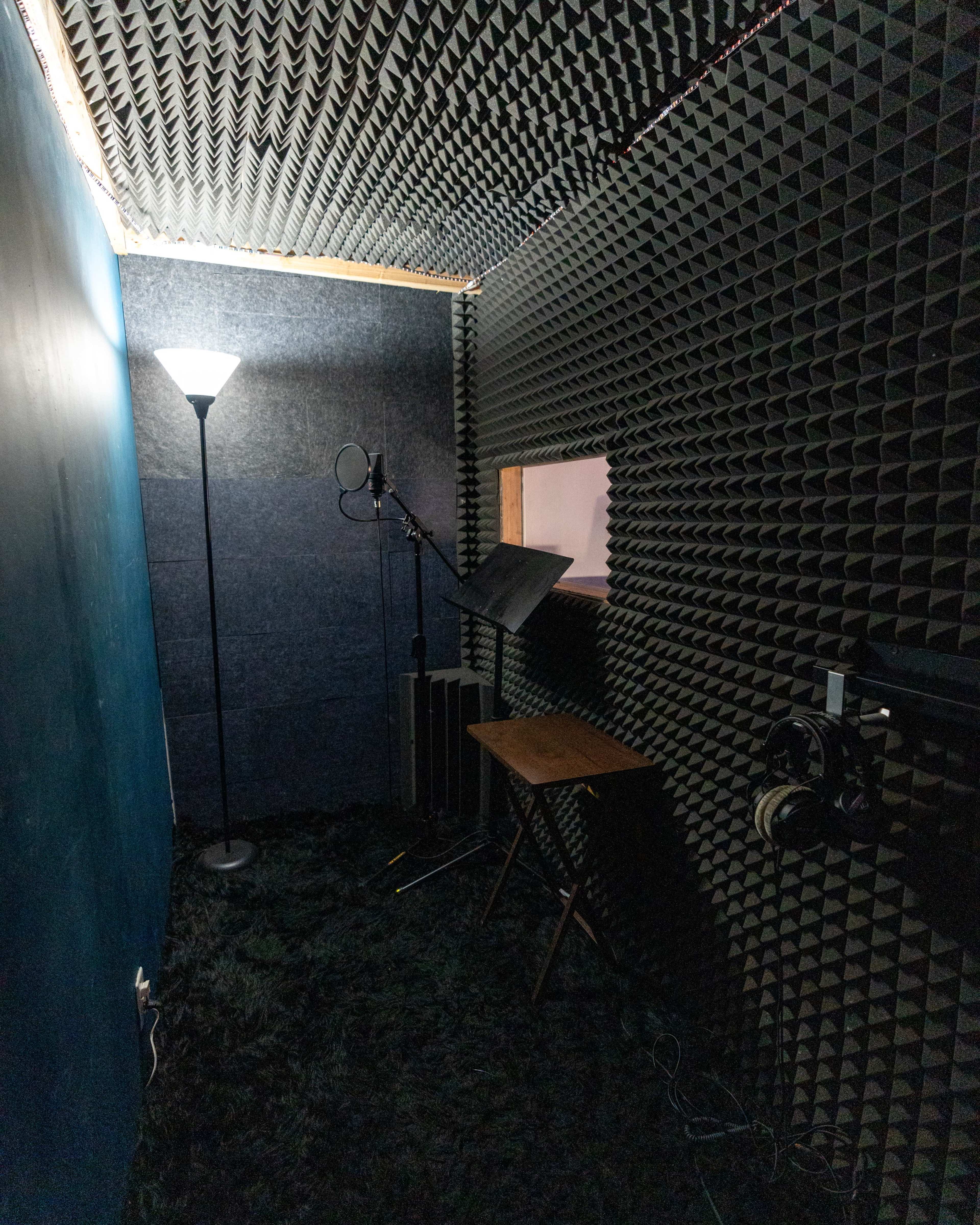 The image shows a soundproofed recording studio with acoustic foam on the walls, a microphone stand, a music stand, a floor lamp, and a small table.