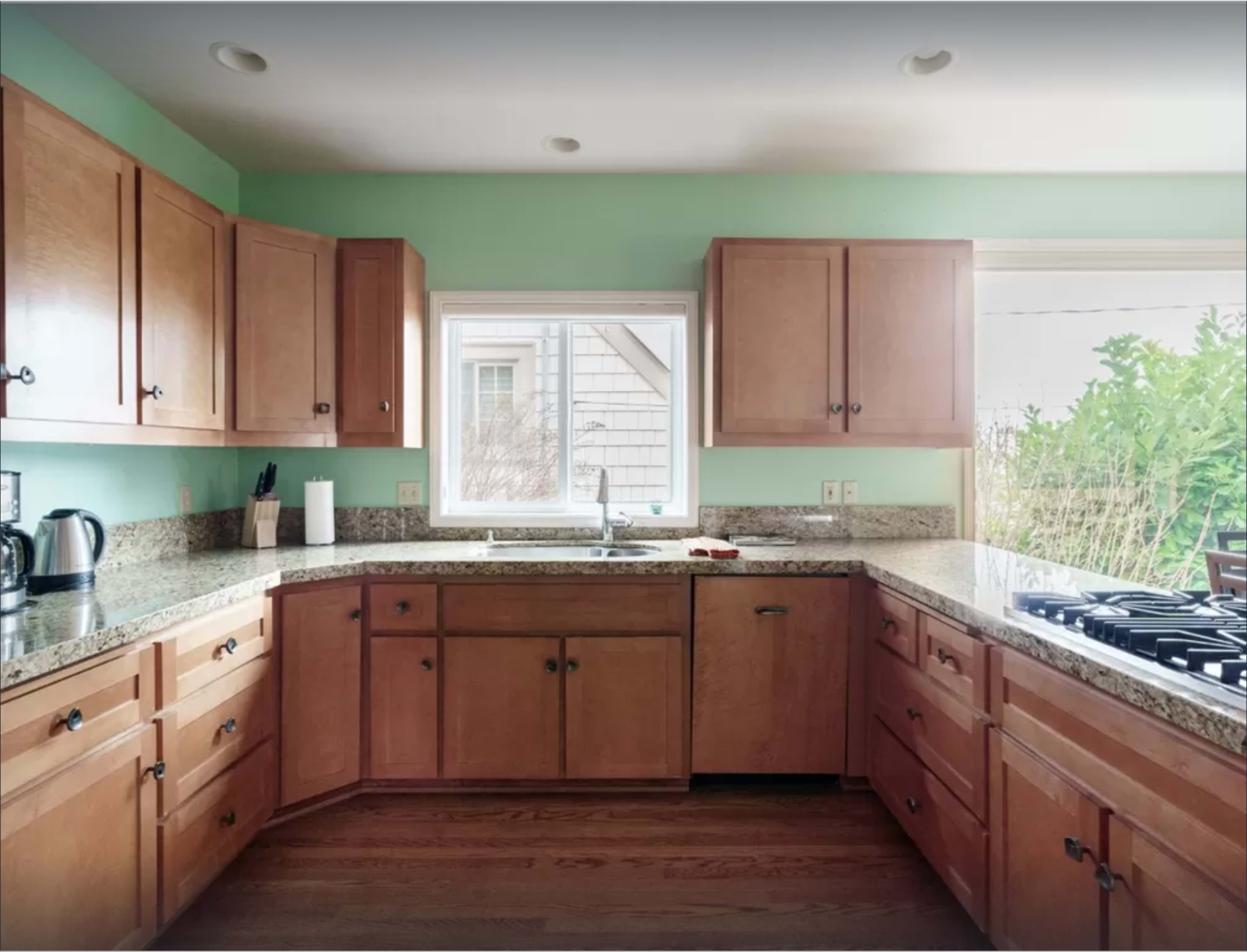 A well-lit kitchen features wooden cabinets, a granite countertop, and a gas stove near a window overlooking greenery.