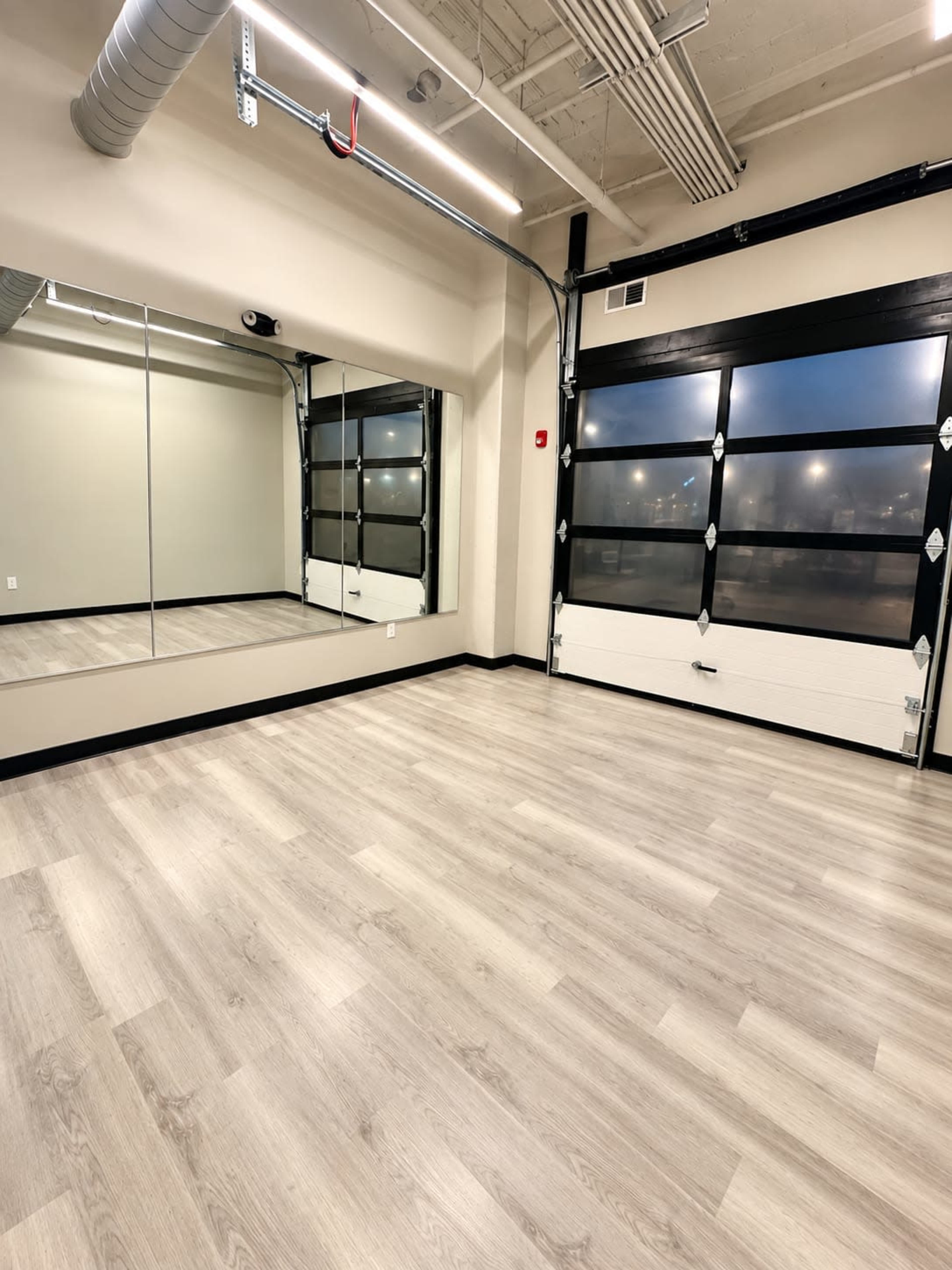 The image shows an empty room with a wood-paneled floor, a wall of mirrors, and a large garage-style door partially lit by external light.