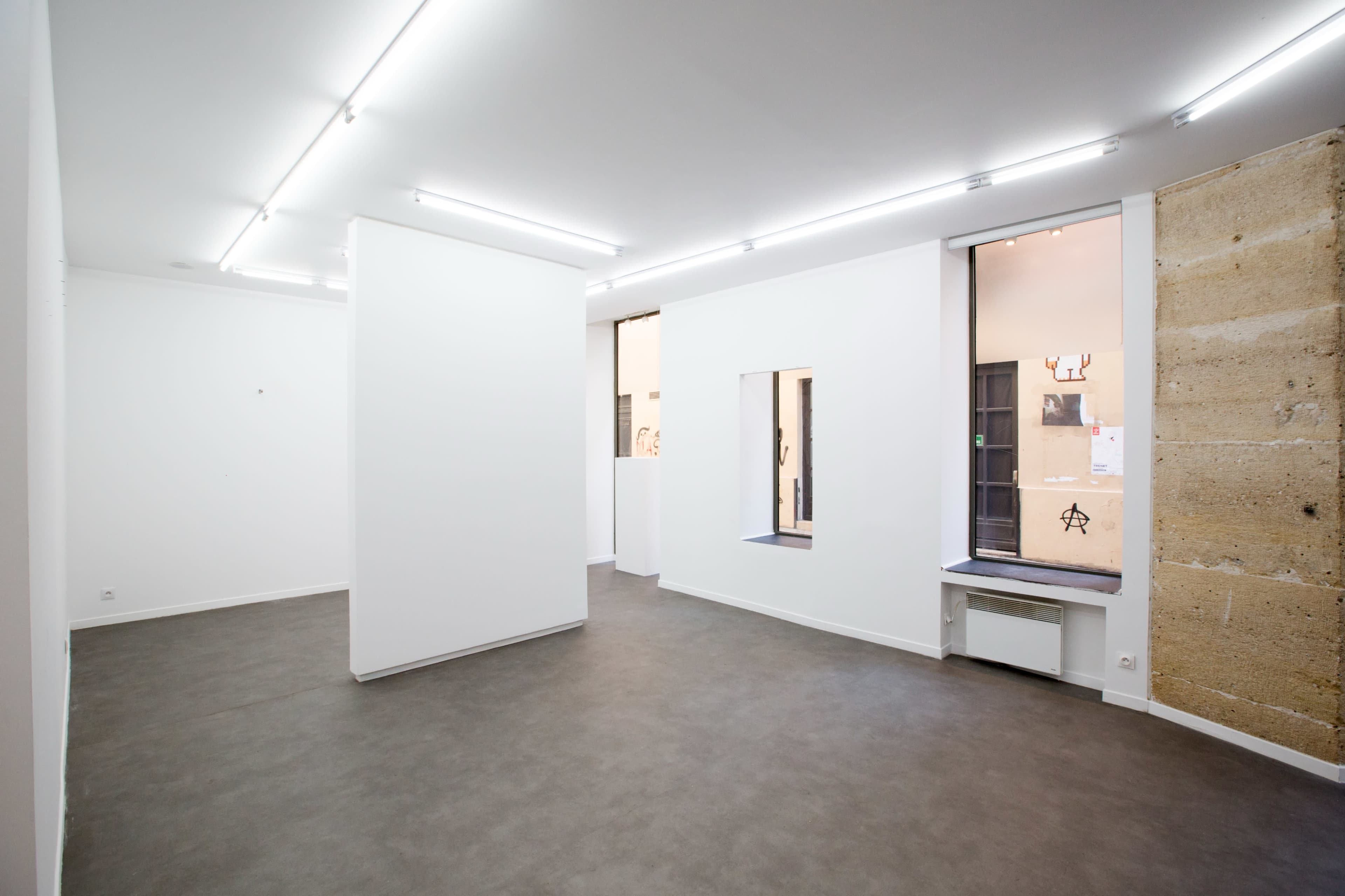 The image shows a minimalist gallery space with white walls, a concrete floor, and large windows providing natural light.