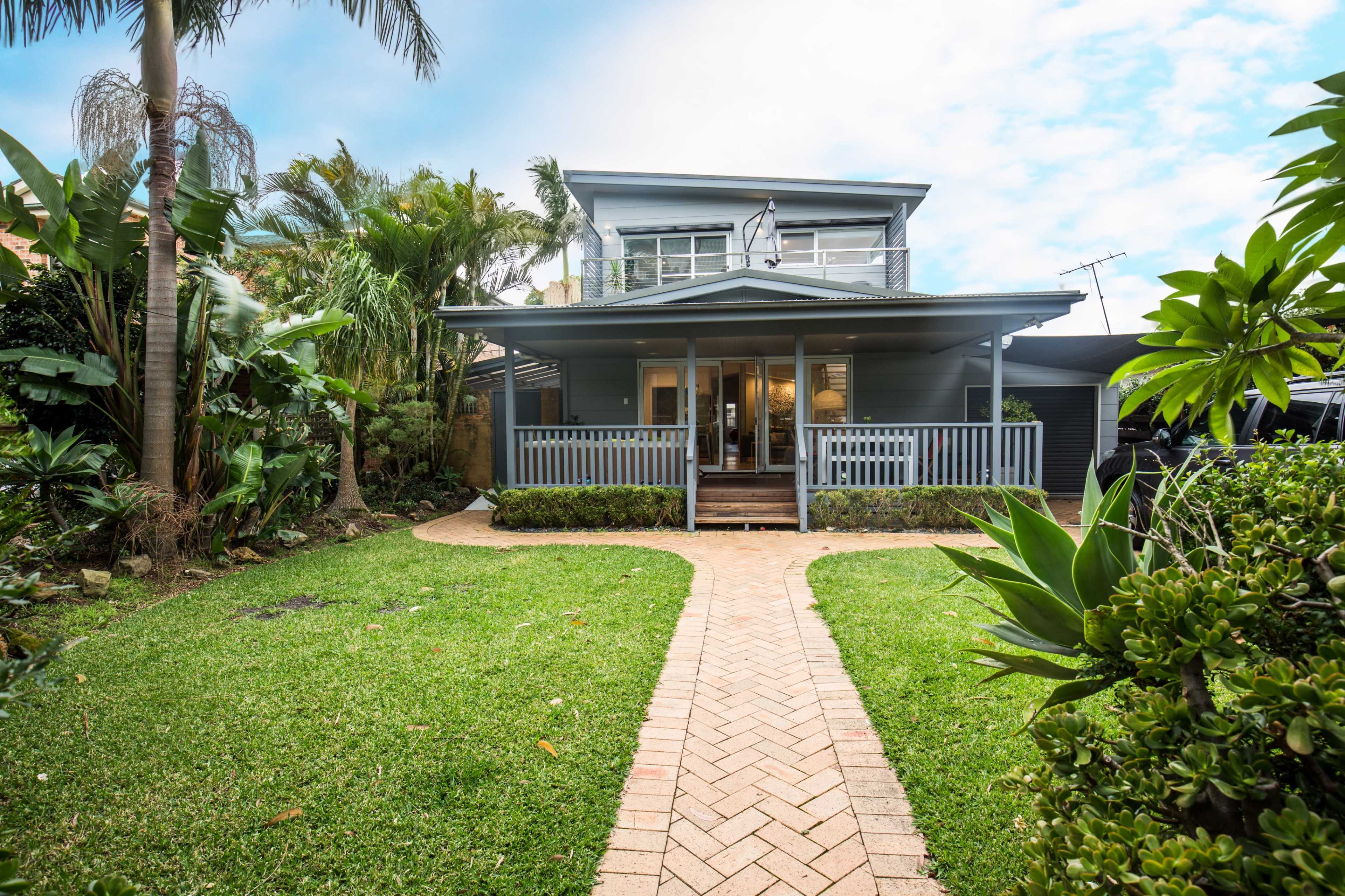 The image shows a two-story house surrounded by lush greenery and a brick pathway leading to the entrance.