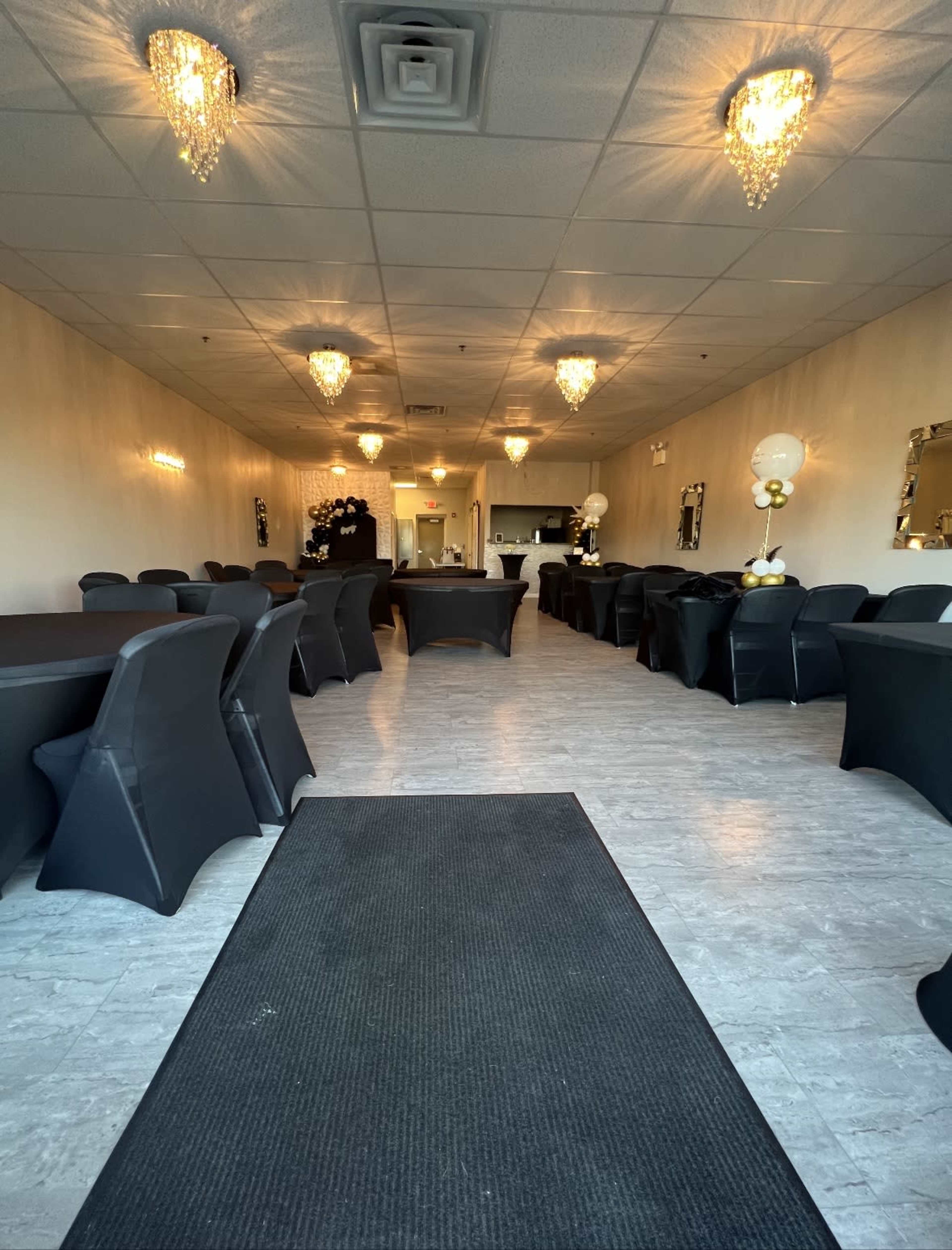 A spacious event venue with black tablecloths on round tables, adorned with white and gold decorations, and elegant chandeliers hanging from the ceiling.