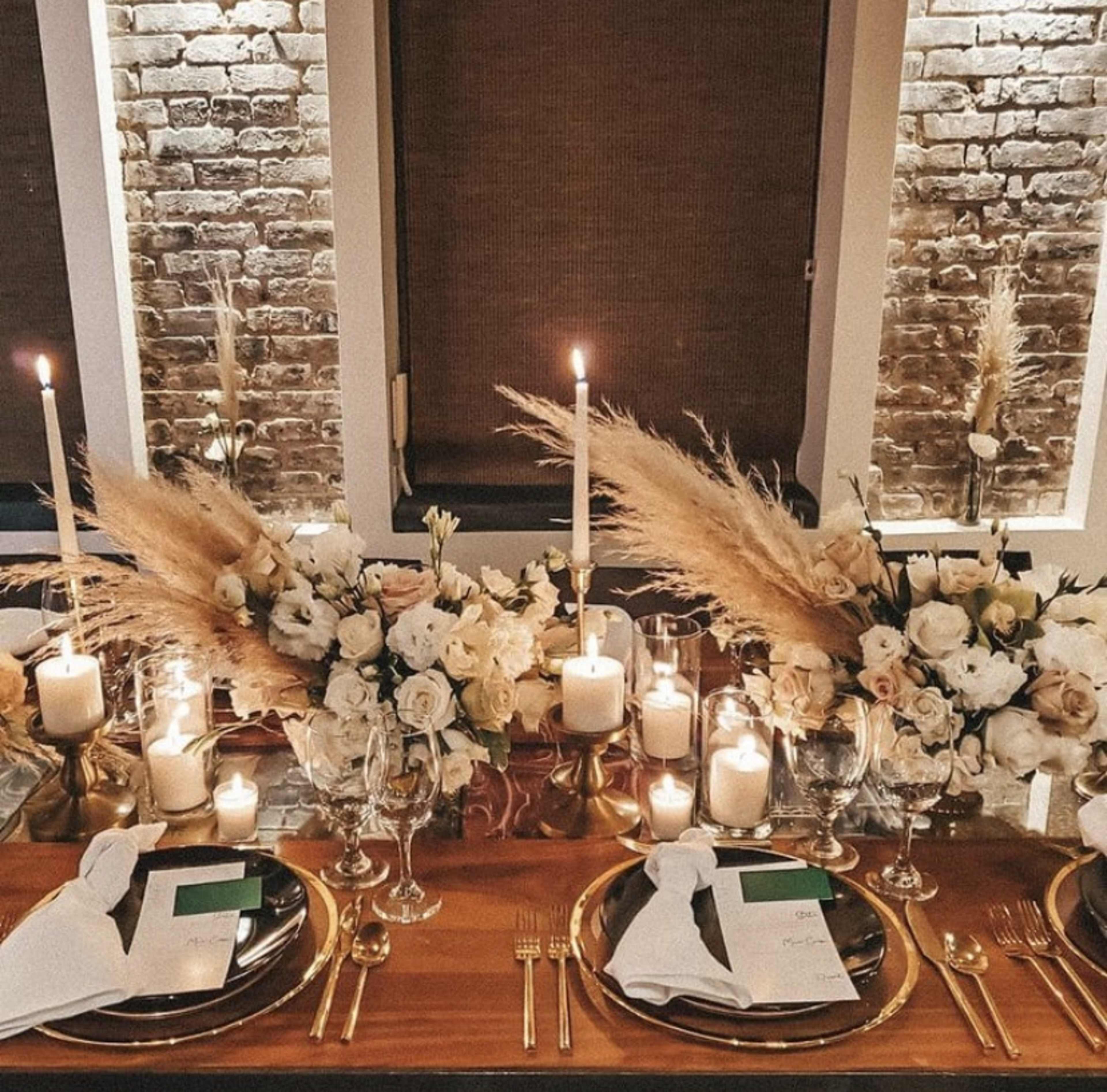 A beautifully arranged dining table features elegant place settings, tall candles, and floral centerpieces against a rustic brick backdrop.
