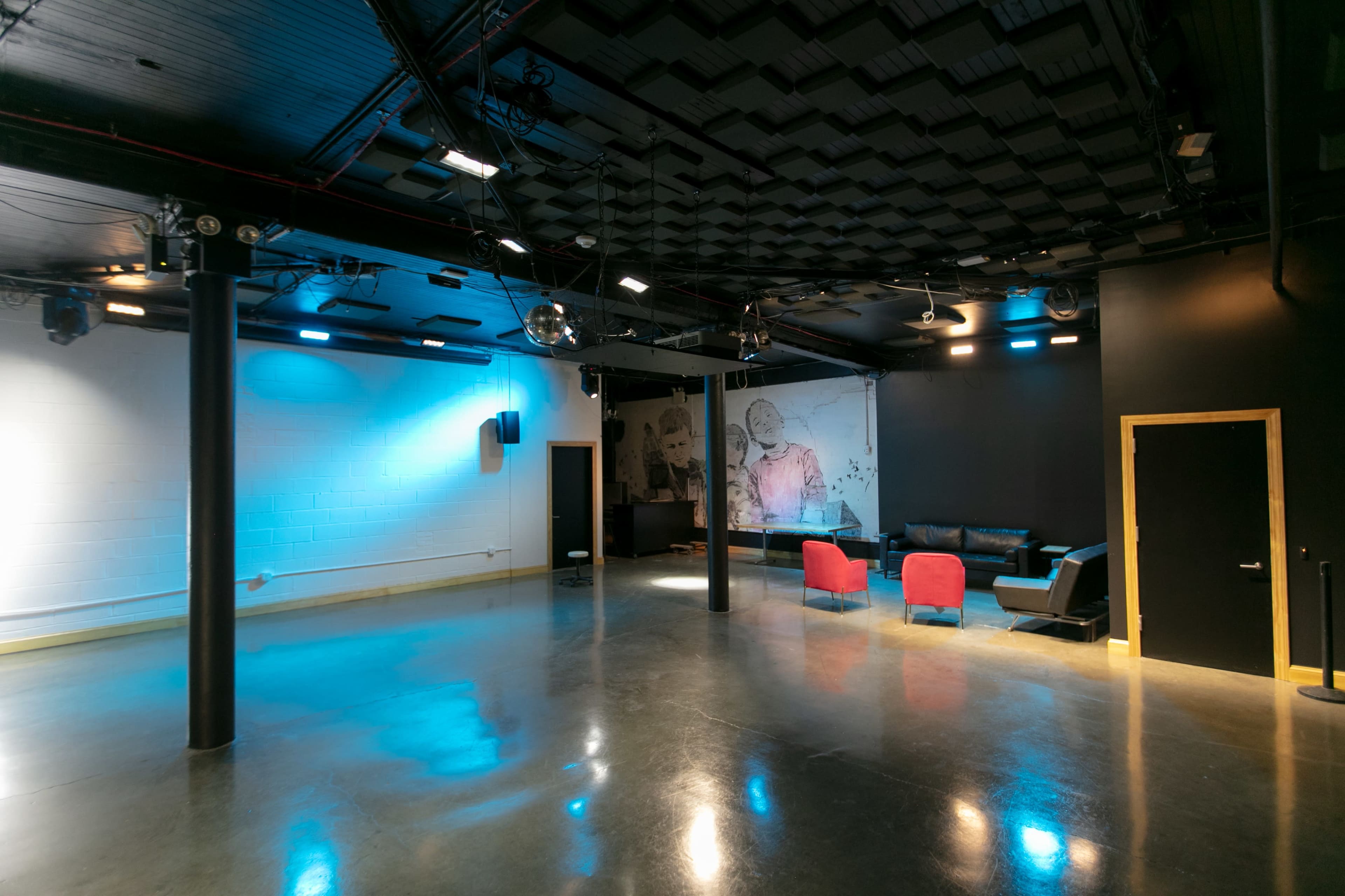 A spacious room with polished concrete floors, black walls, and minimal seating consisting of a black couch and two red chairs, illuminated by blue and white lighting.