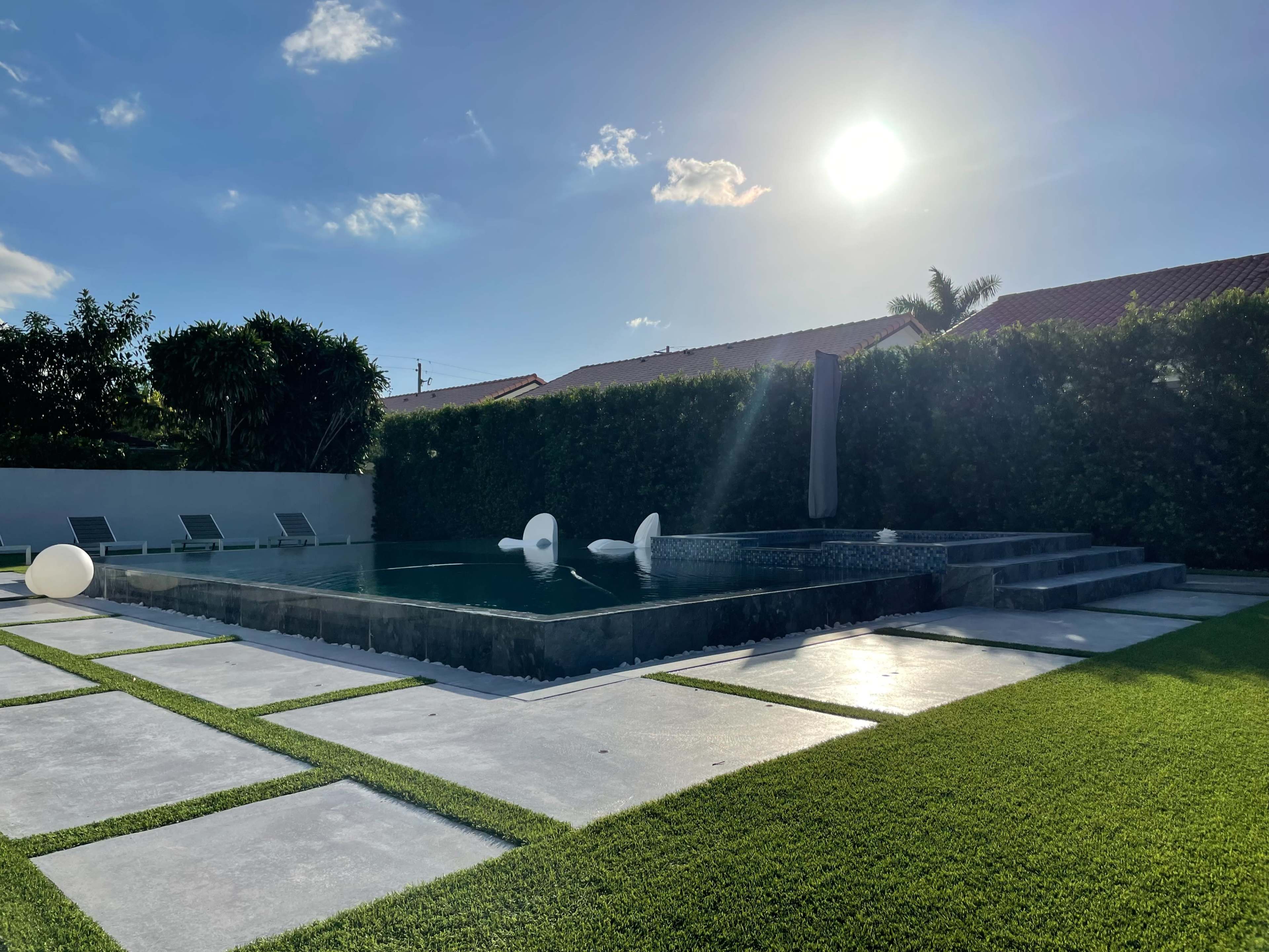 The image shows a modern swimming pool surrounded by lush greenery and a well-maintained lawn under a bright sun.