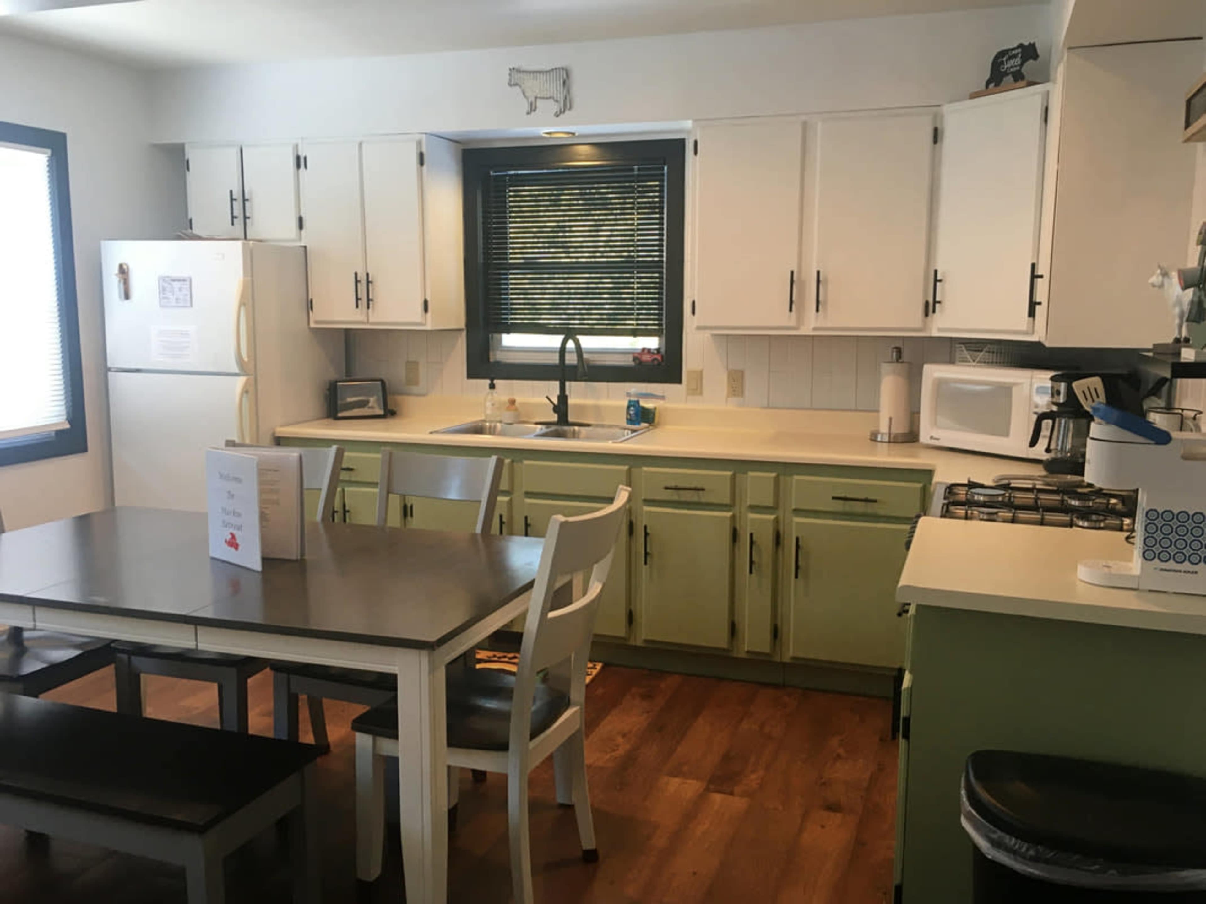 A kitchen features white cabinets with green accents, a table with chairs, a refrigerator, a microwave, and a gas stove.